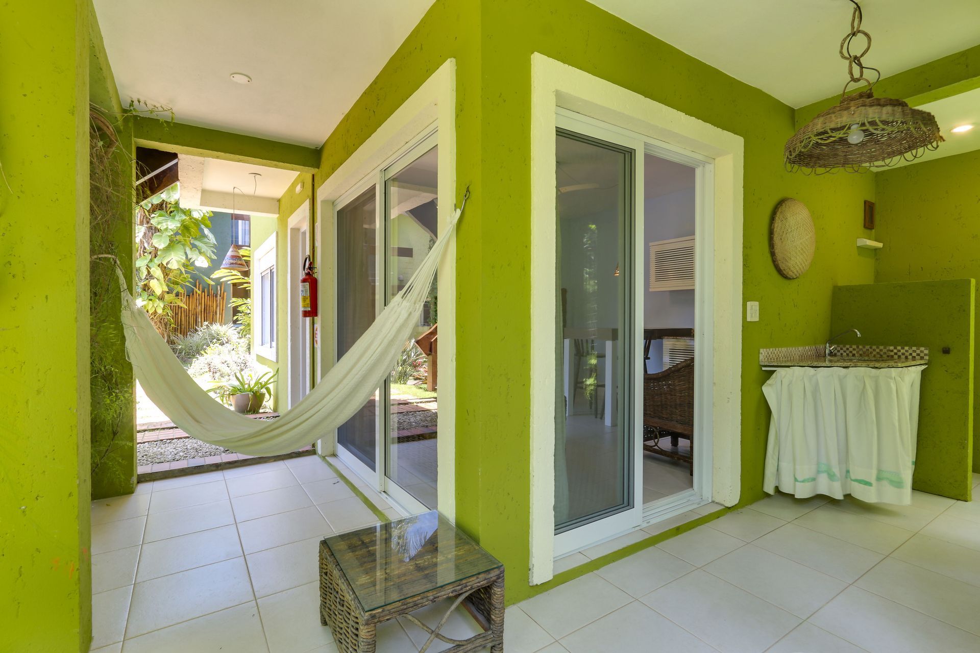 Pátio verde-limão vibrante com rede branca, portas de vidro e corredor com piso de azulejo.