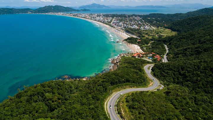 Vista aérea de uma estrada costeira serpenteando ao longo de uma baía turquesa e colinas verdejantes perto de uma cidade litorânea.