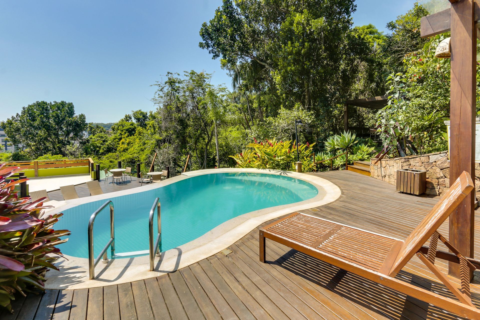 Piscina no quintal com deck de madeira, espreguiçadeira e árvores verdejantes sob a luz do sol.