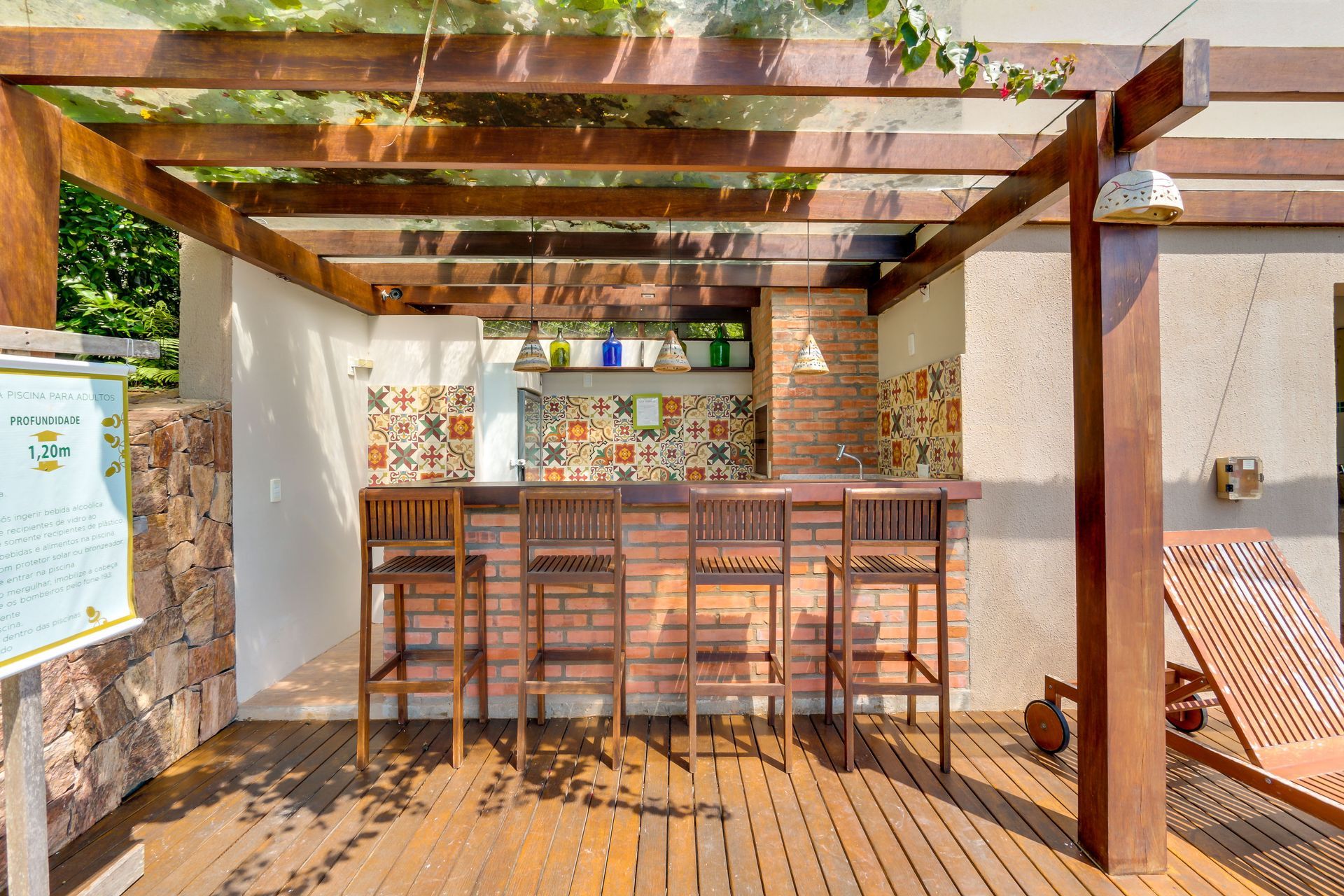 Área de bar externa com banquetas de madeira, pérgola e balcão de azulejos em um pátio ensolarado.