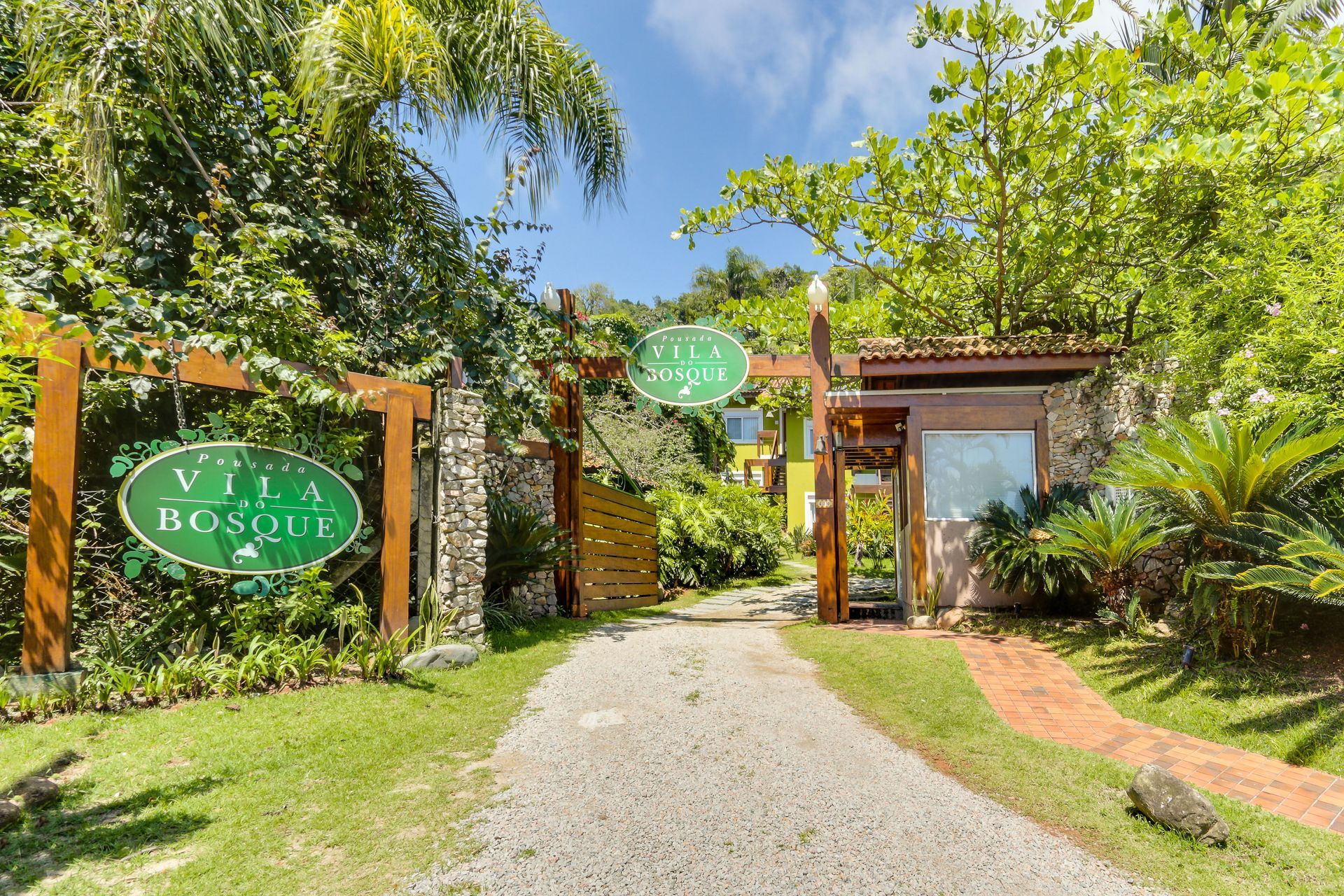 Entrada da Villa Bosco com caminho de cascalho, portão de madeira e paisagismo tropical exuberante.