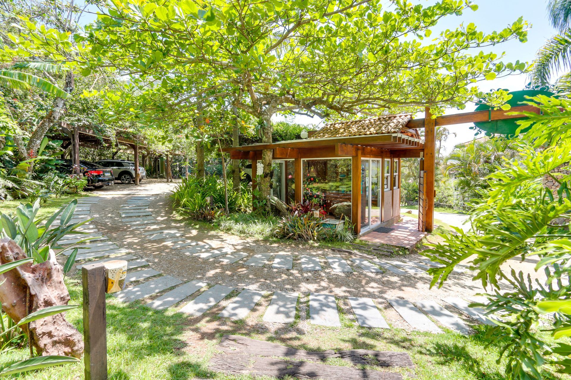 Caminho sombreado no jardim, ao lado de um pavilhão de madeira, com árvores verdejantes e muita luz solar.