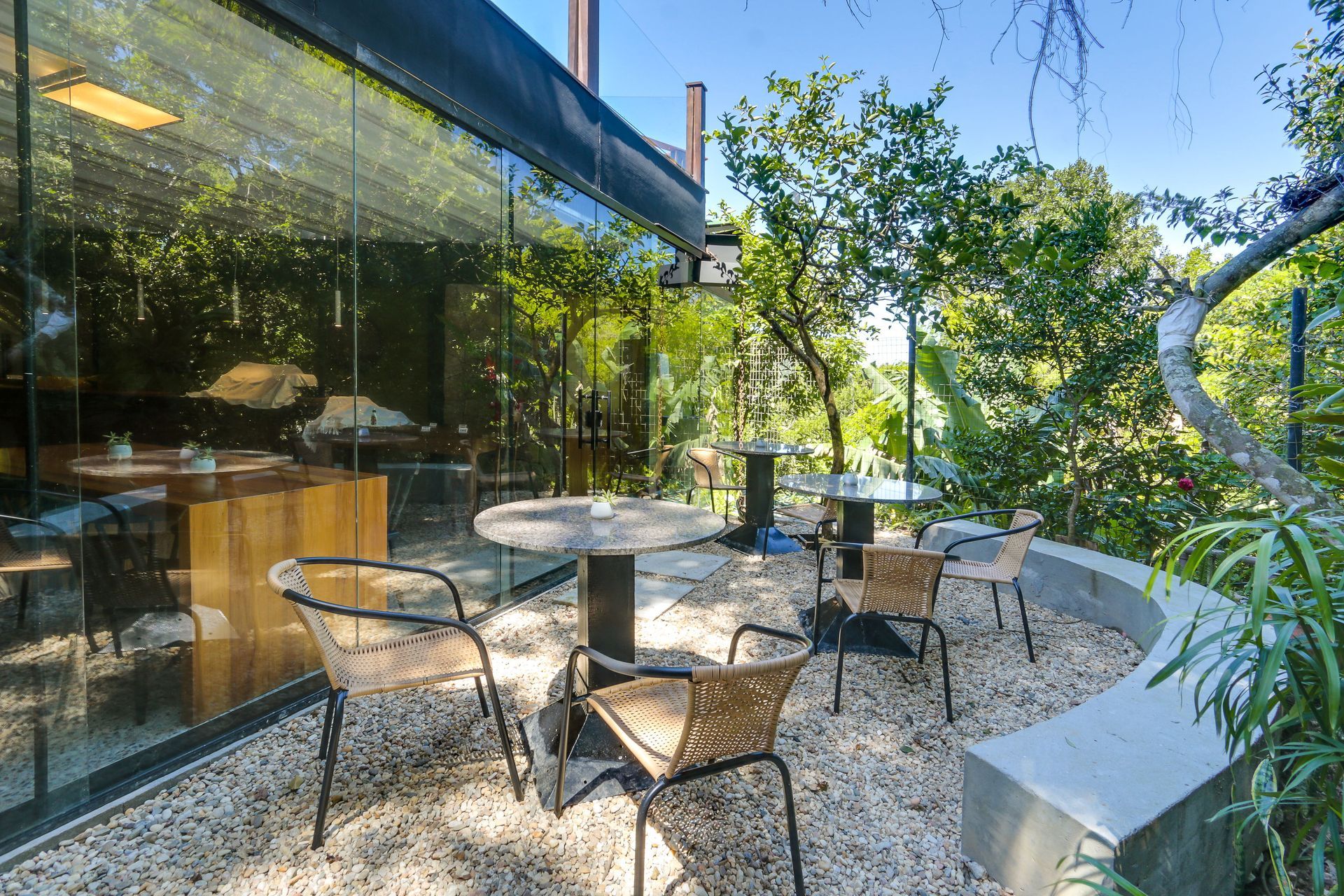 Pátio externo com cadeiras e mesas de metal ao lado de um edifício com paredes de vidro, sob árvores.