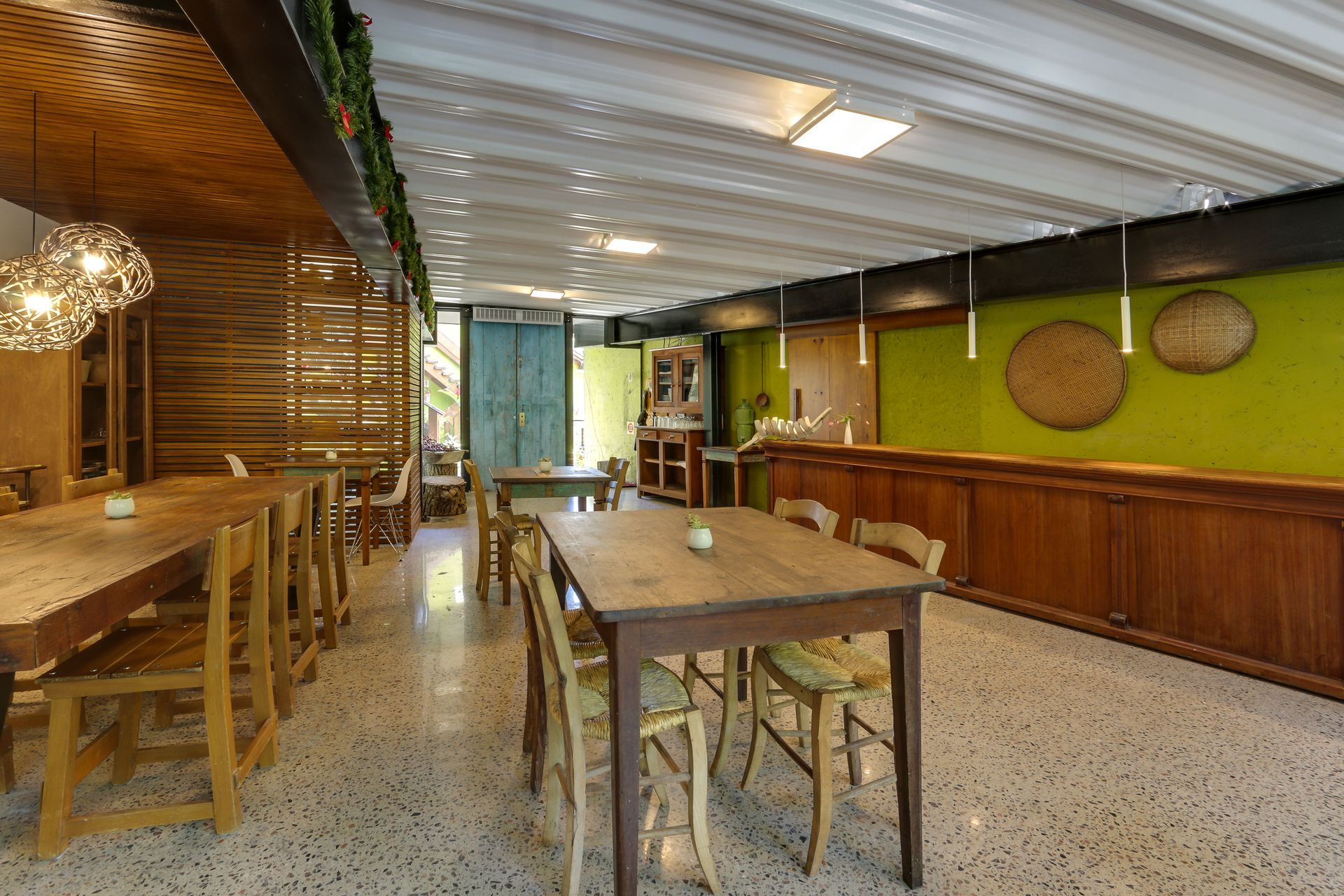Café rústico vazio com mesas de madeira, cadeiras de vime e uma parede verde com cestos suspensos.