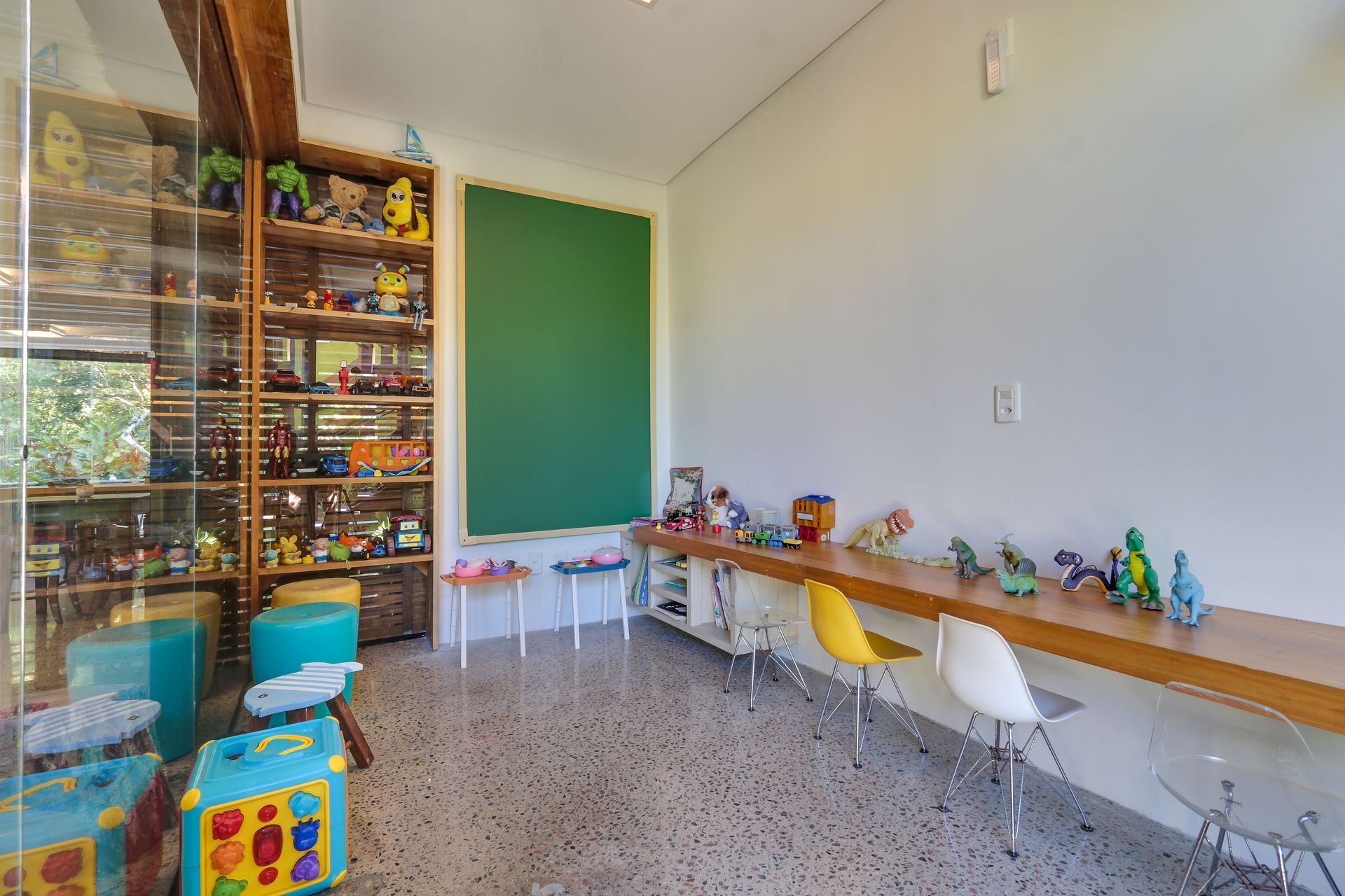 Canto da sala de aula com prateleiras, brinquedos, cadeirinhas e uma longa mesa de madeira encostada em uma parede branca.