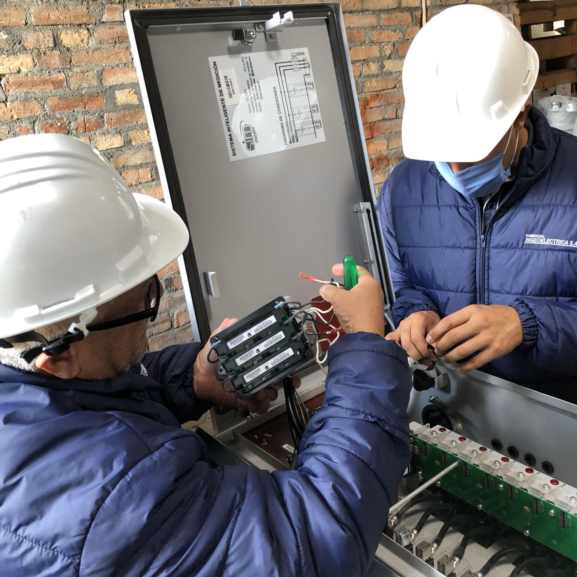 Dos hombres con cascos y máscaras están trabajando en una caja eléctrica.