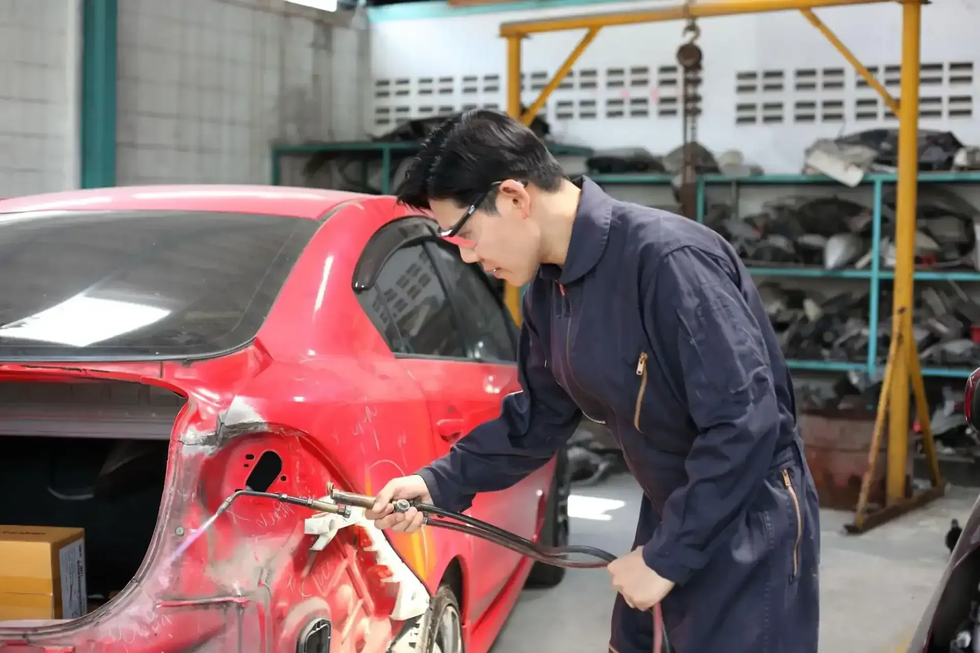 A mechanic in dark coveralls uses a welding torch to repair the rear bodywork