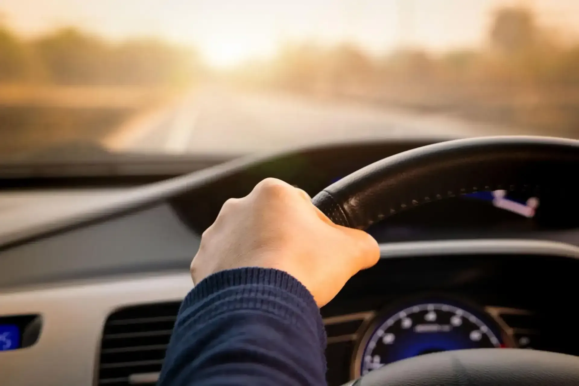 A person’s hand grips a car steering wheel as they drive down a road toward a bright sunset.