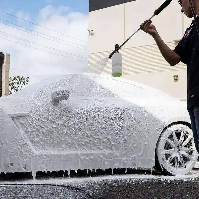 A person uses a pressure washer with a foam cannon to apply thick white soap to the side of a car.