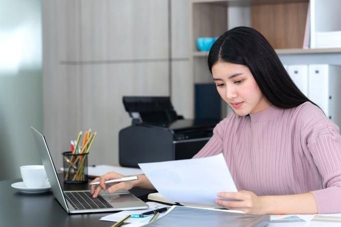 A person in a pink sweater sits at a desk, typing on a laptop while holding document.