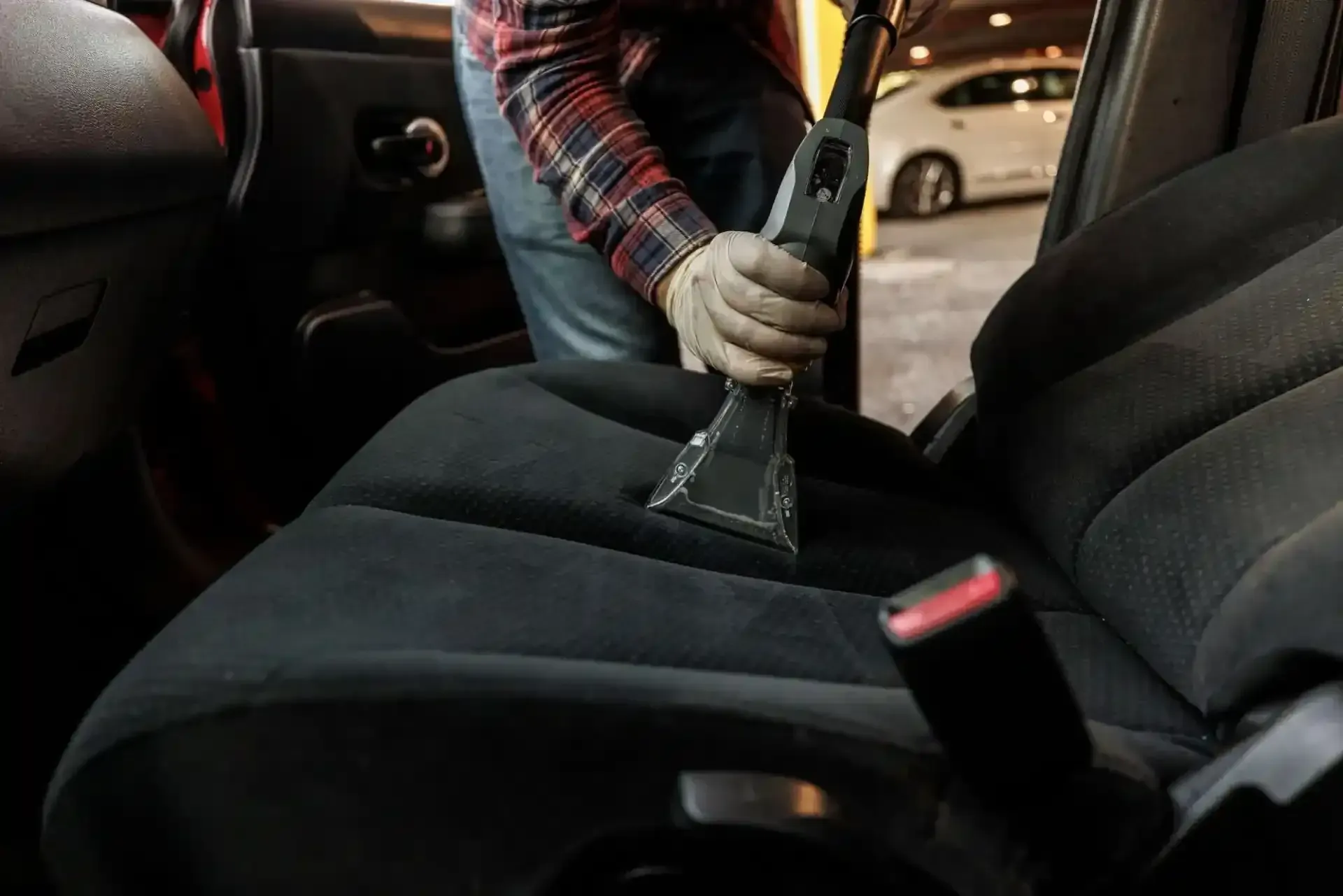 A person uses an upholstery cleaner tool on the black fabric seat of a car interior.