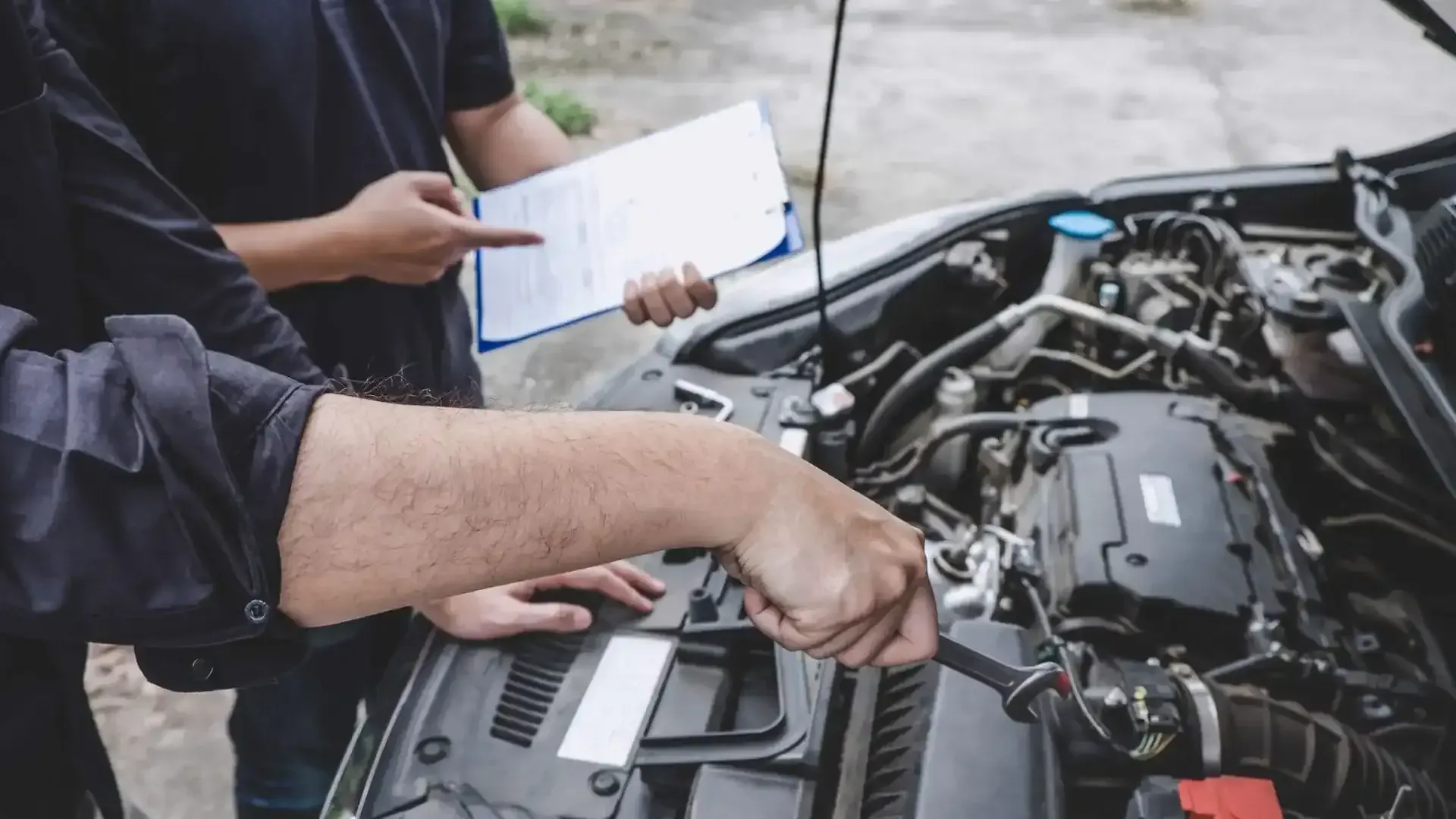 Two people in dark work shirts inspect a car engine; one points to a clipboard