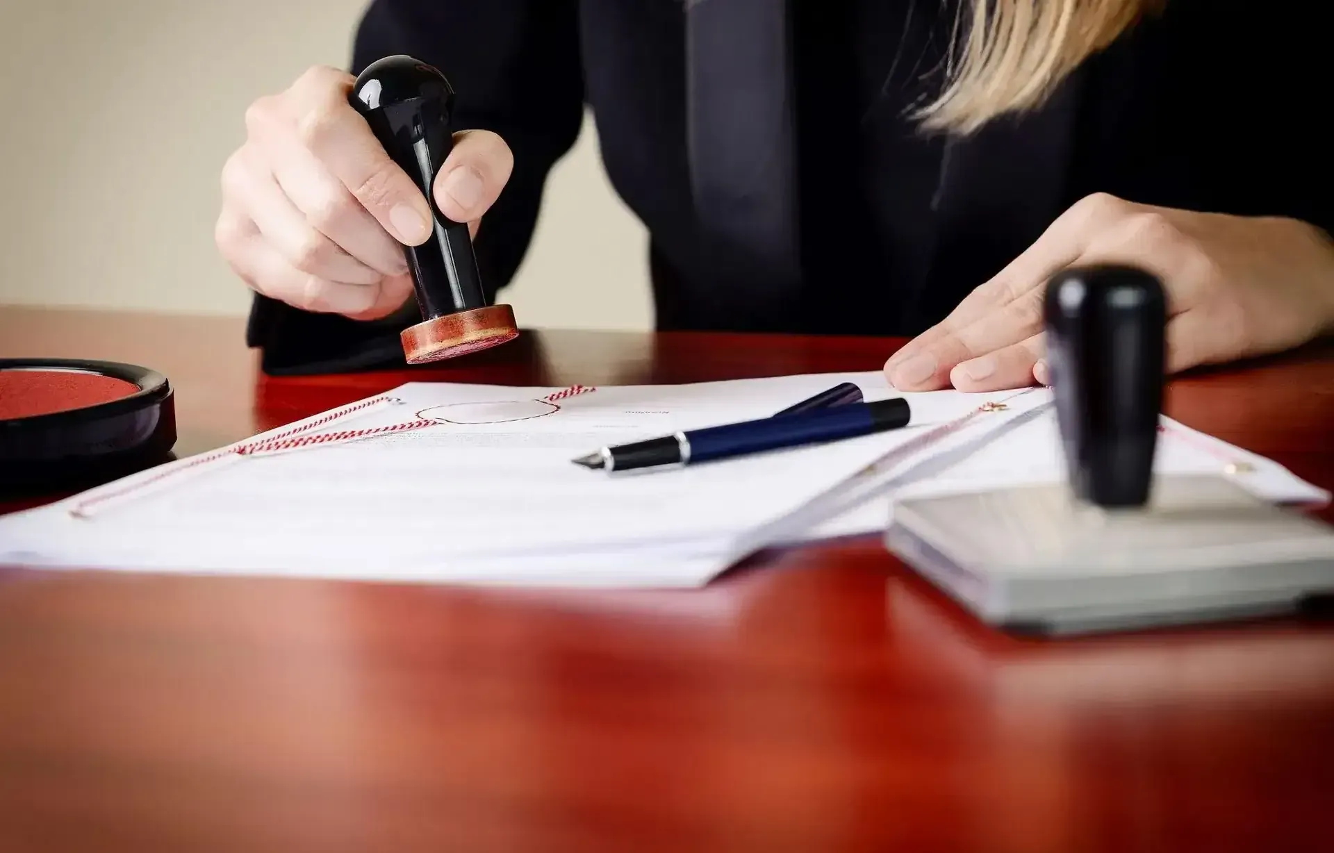 A person in a suit is using a stamp on a document at a wooden desk with a pen