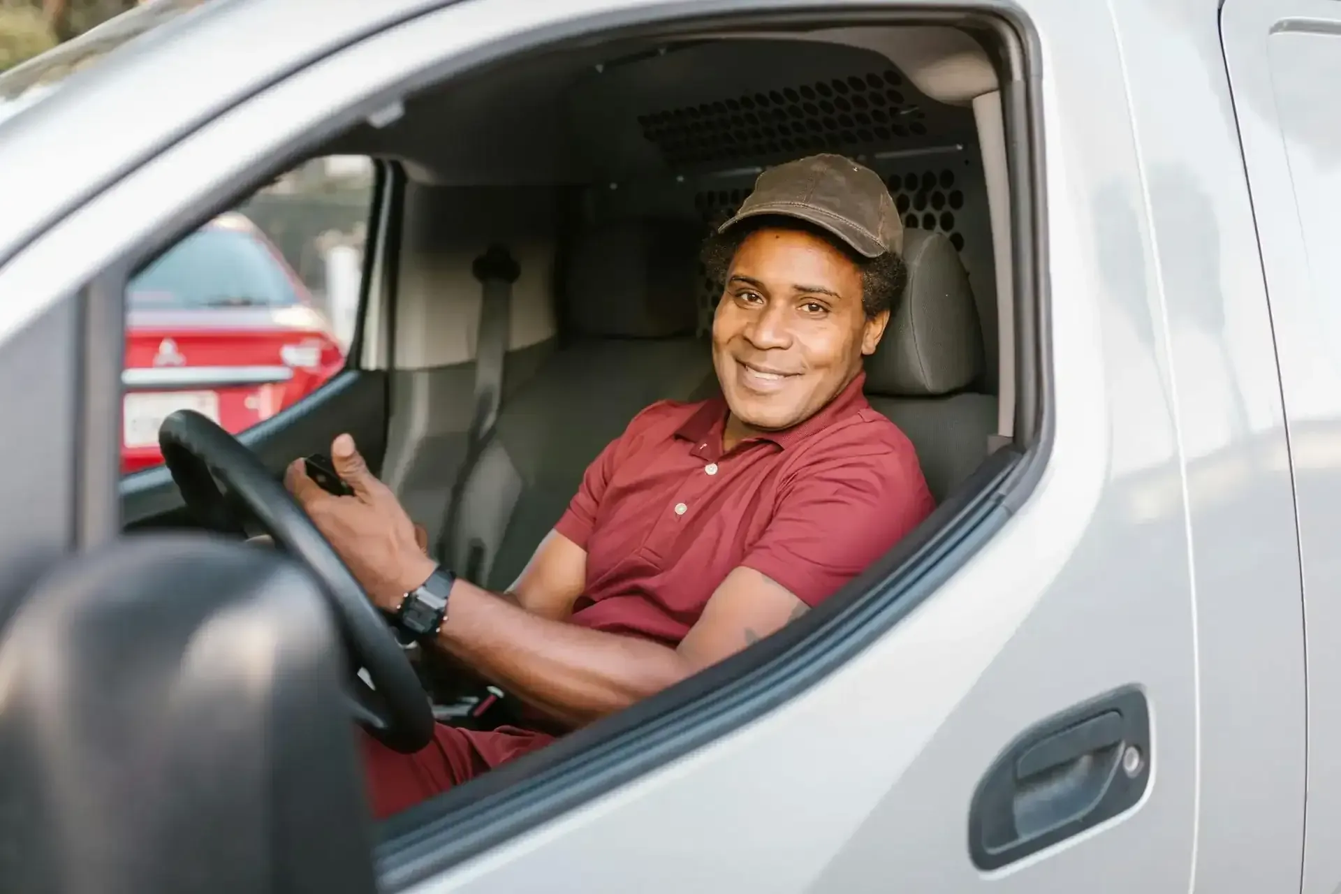 A person in a maroon shirt and cap smiling while driving a white delivery van.