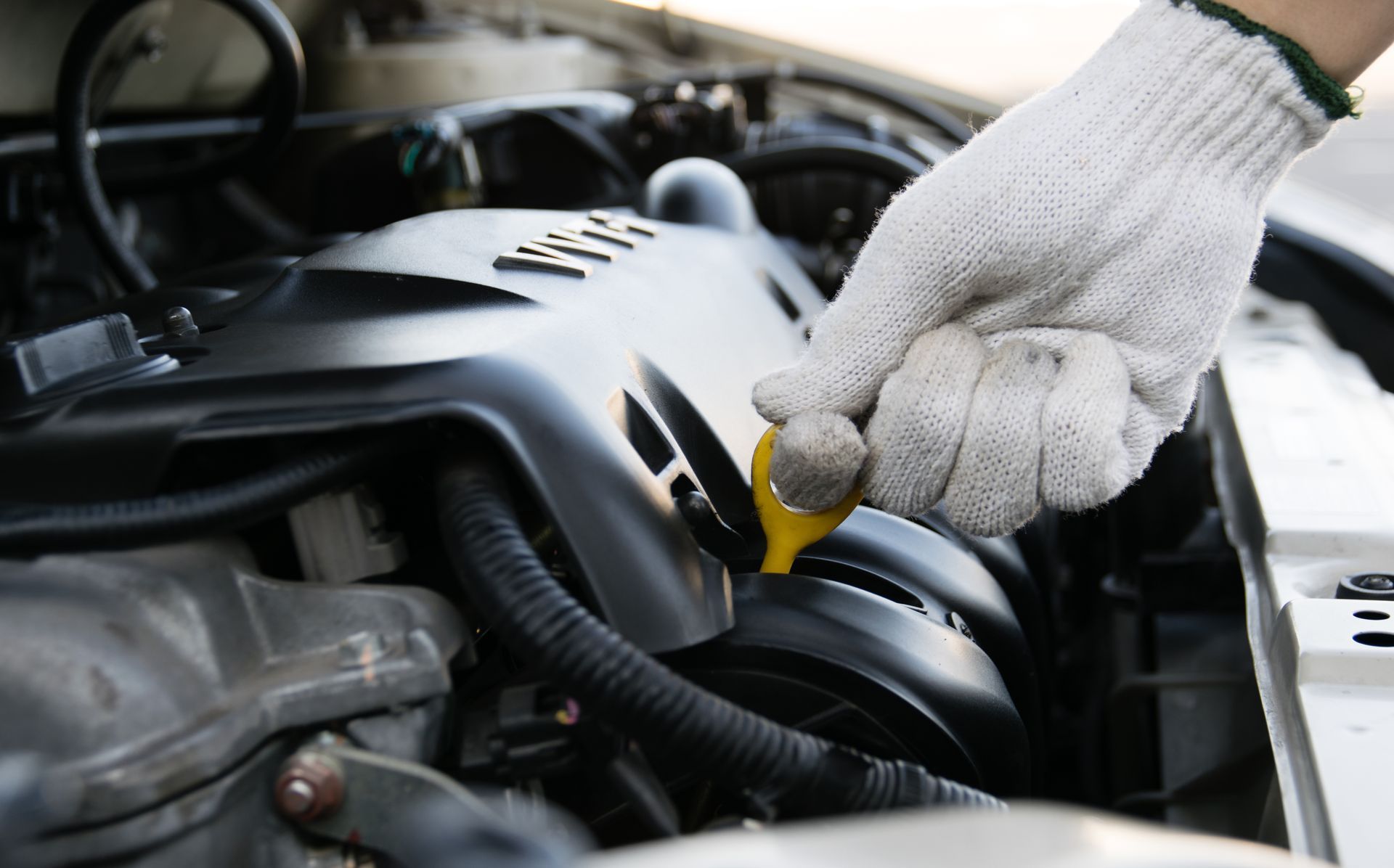 A person wearing work gloves checks a car engine's oil level by pulling the yellow dipstick 