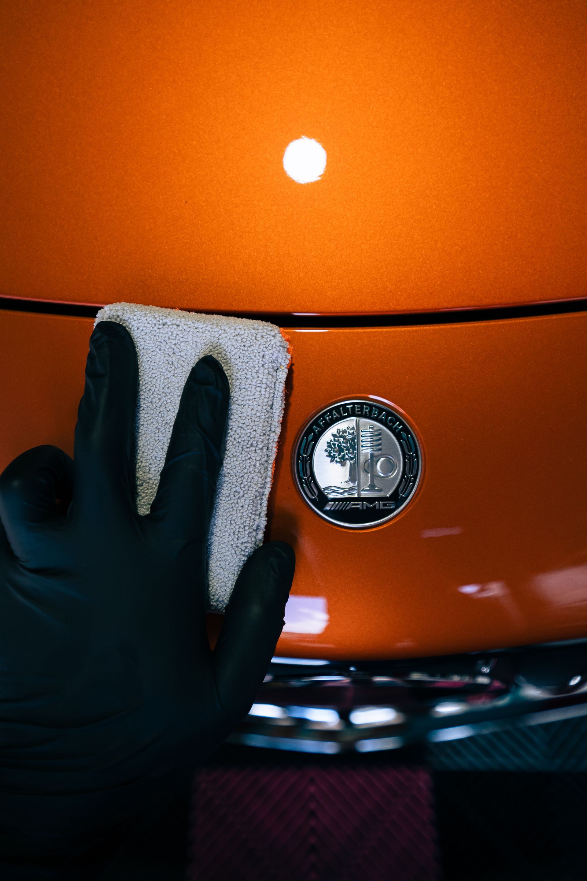 Gloved hand applying ceramic coating on an orange car, near a chrome AMG emblem.