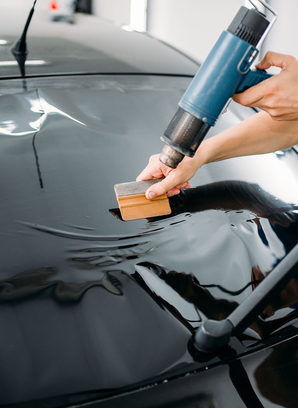 Person applying tint to a car window with a heat gun and squeegee in a shop.