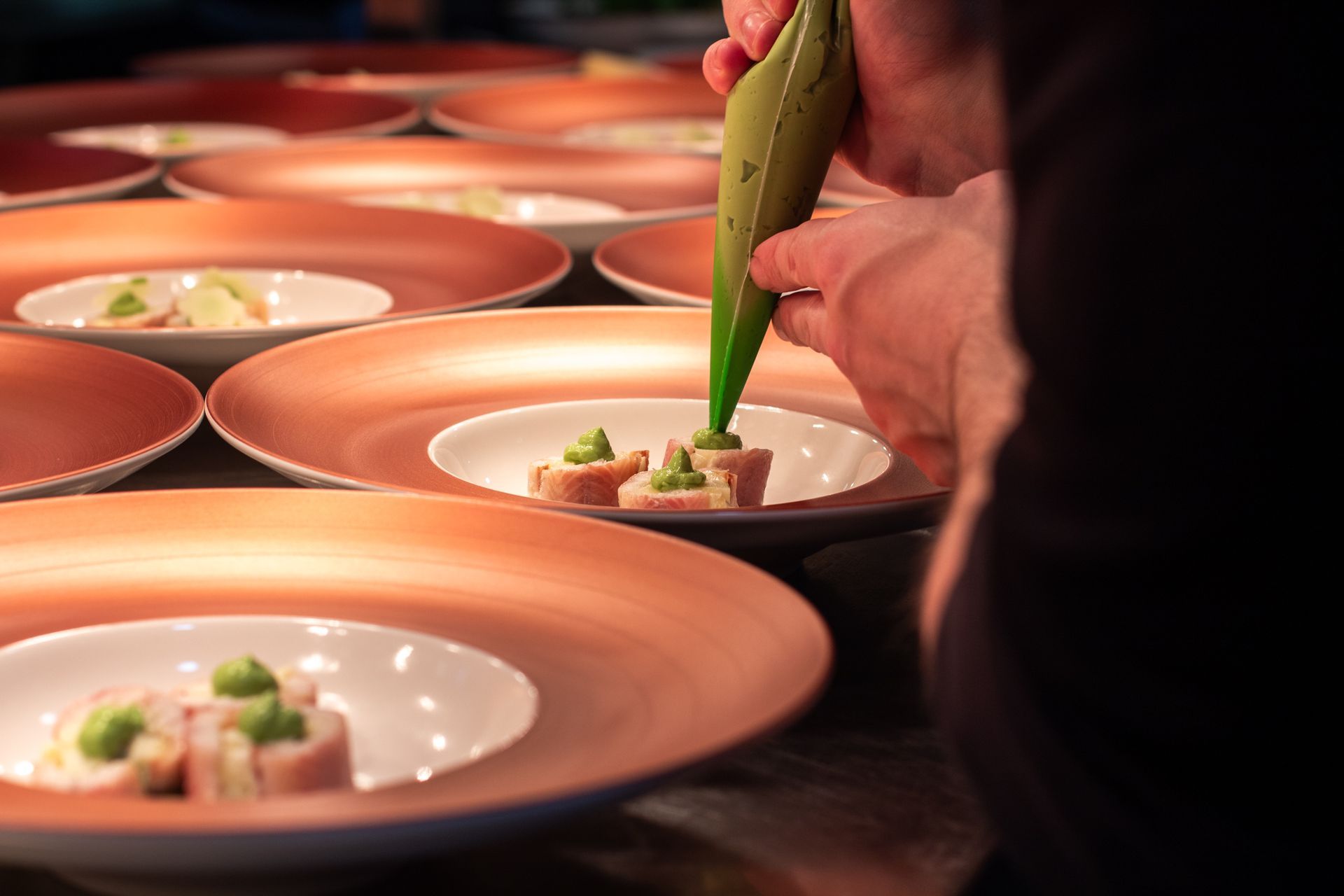 Chef piping green sauce onto plated appetizers on orange-rimmed dishes in a kitchen