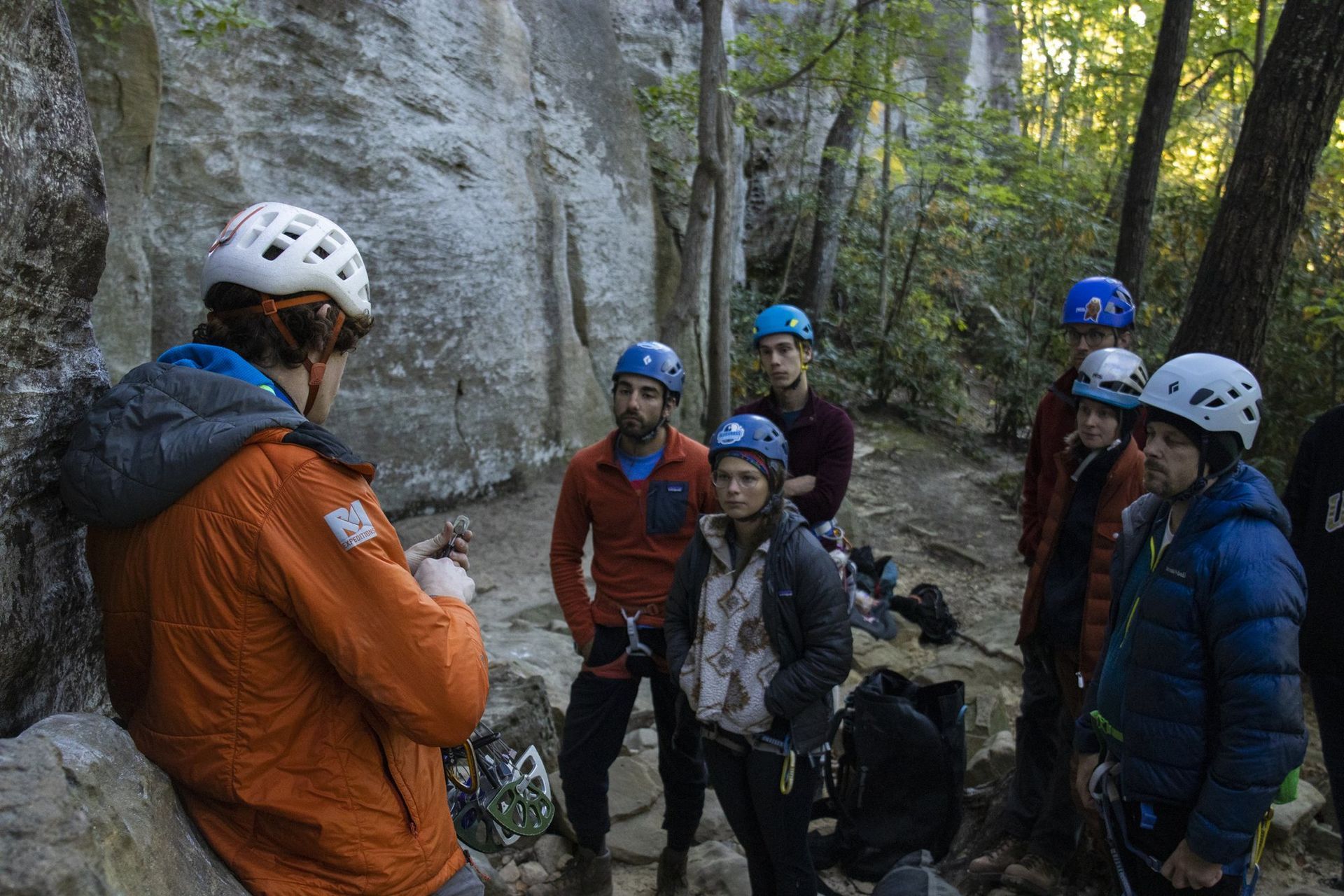 Guide showing climbing gear to a clinic group in helmets at the base of a Red River Gorge cliff