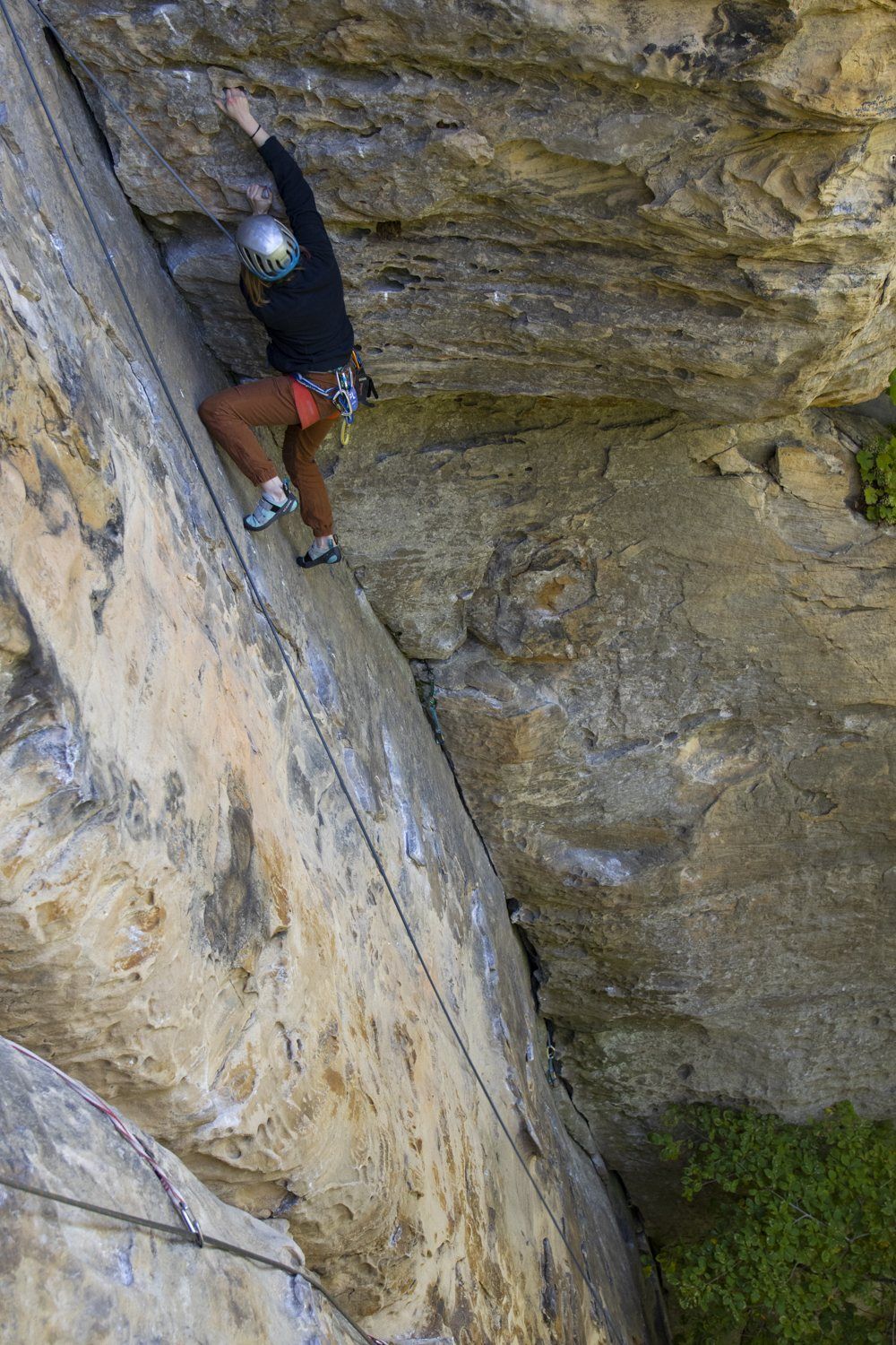 Climber in a helmet moving up a steep Red River Gorge sandstone face below an overhang