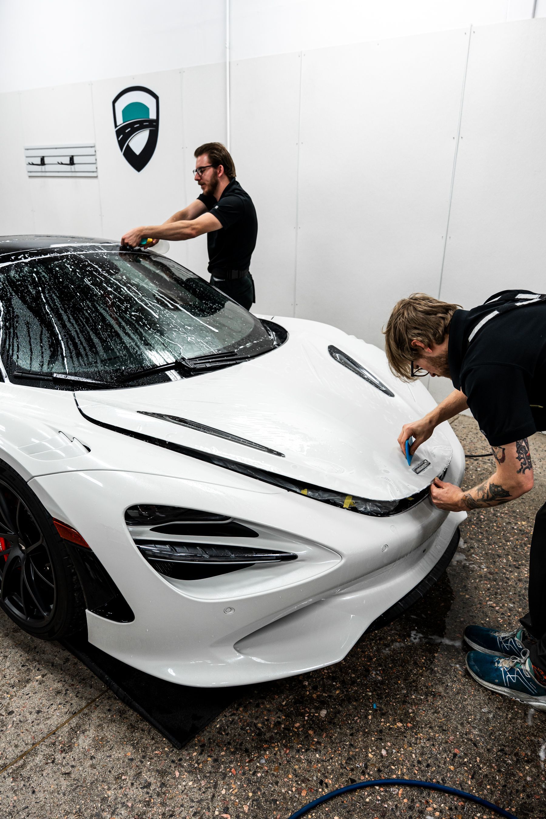 Two men applying protective film to a white McLaren sports car in a workshop.