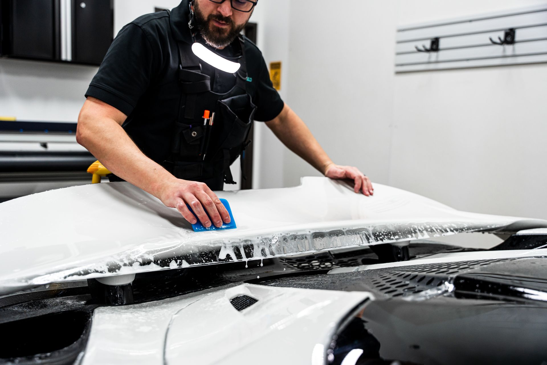 Man applying protective film to a car hood, using a squeegee, in a shop.