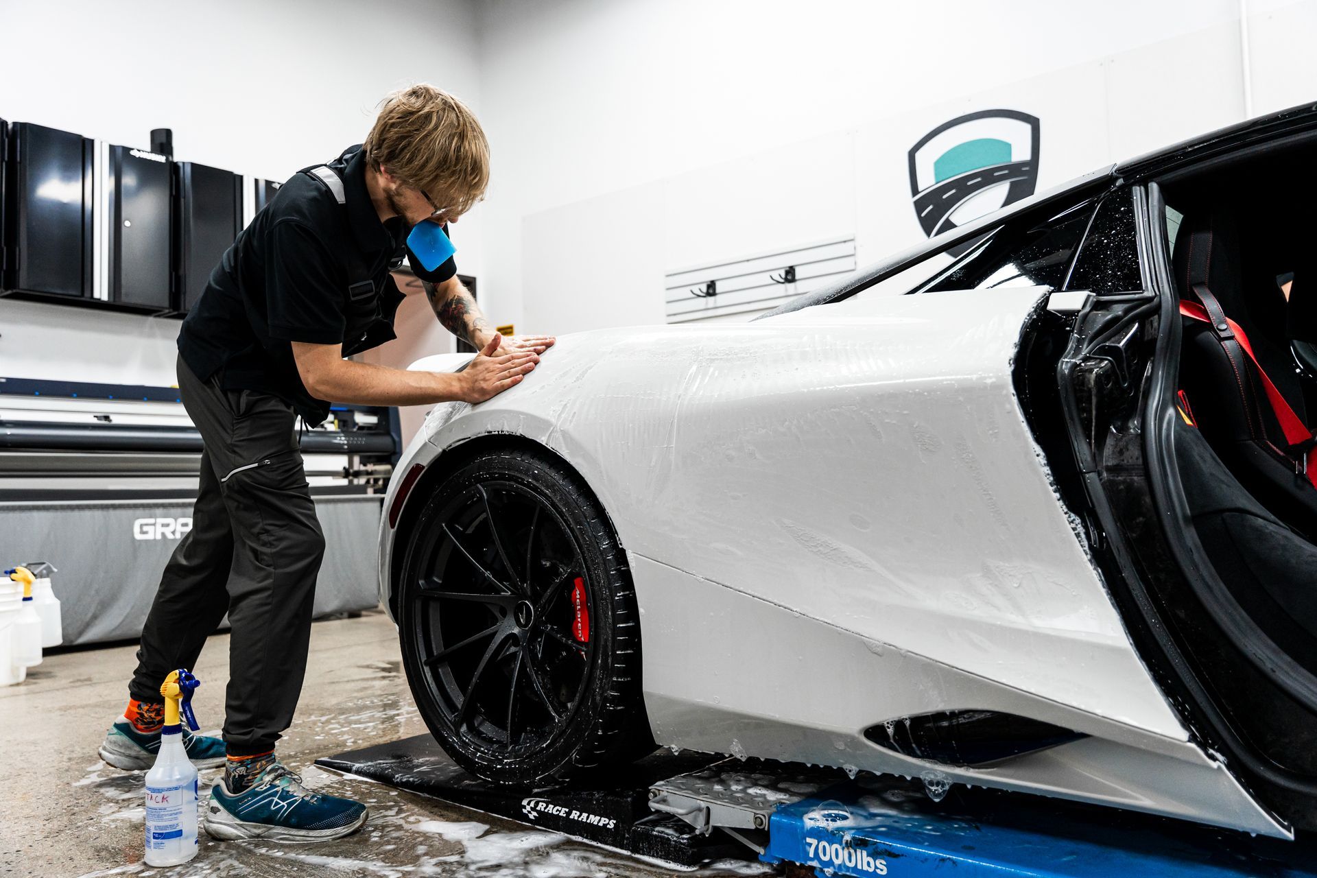 Man applies film to a white car's fender in a shop. He wears a mask and uses soapy water.