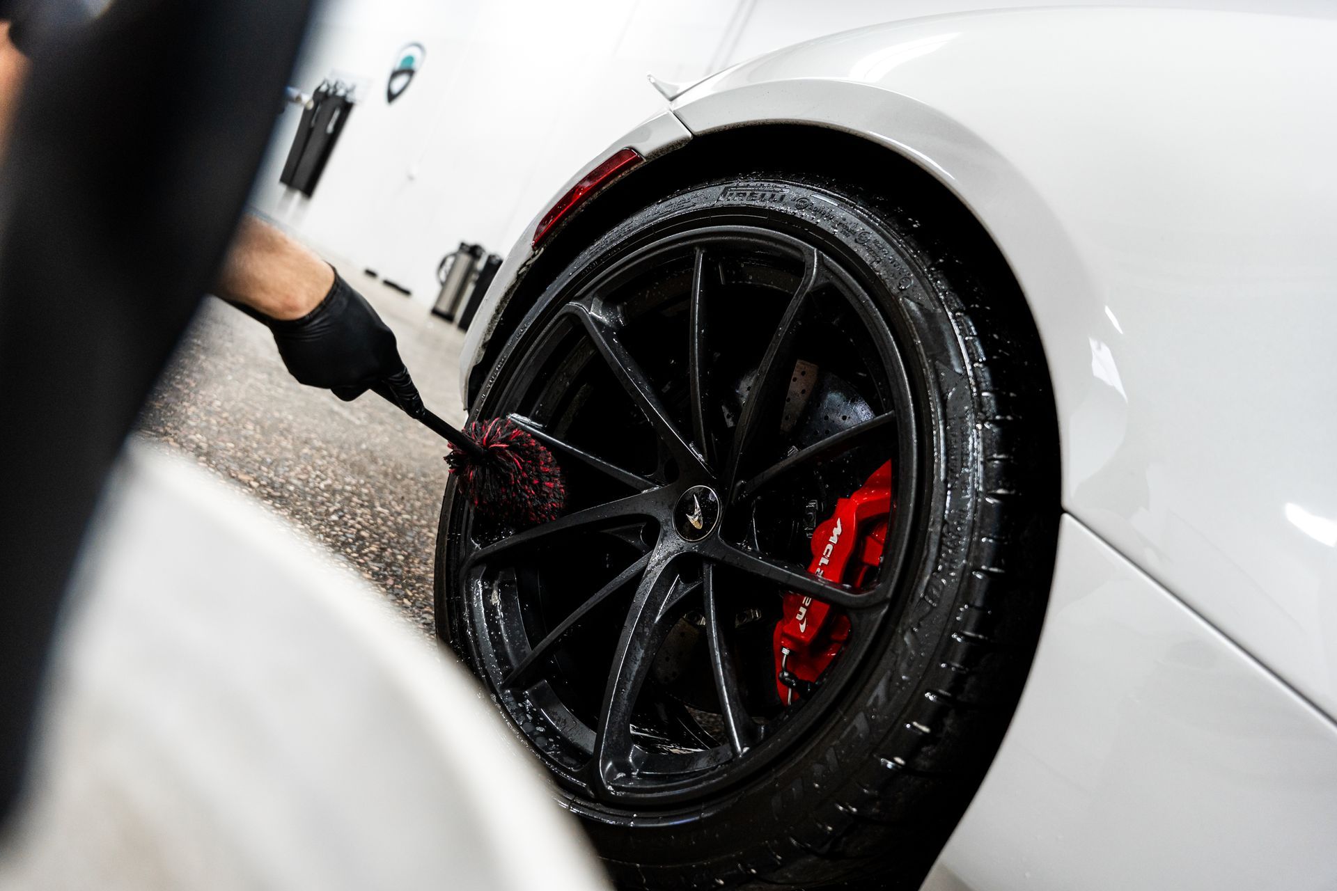 Person wearing black gloves washing a black car wheel with a brush. Red brake caliper visible.