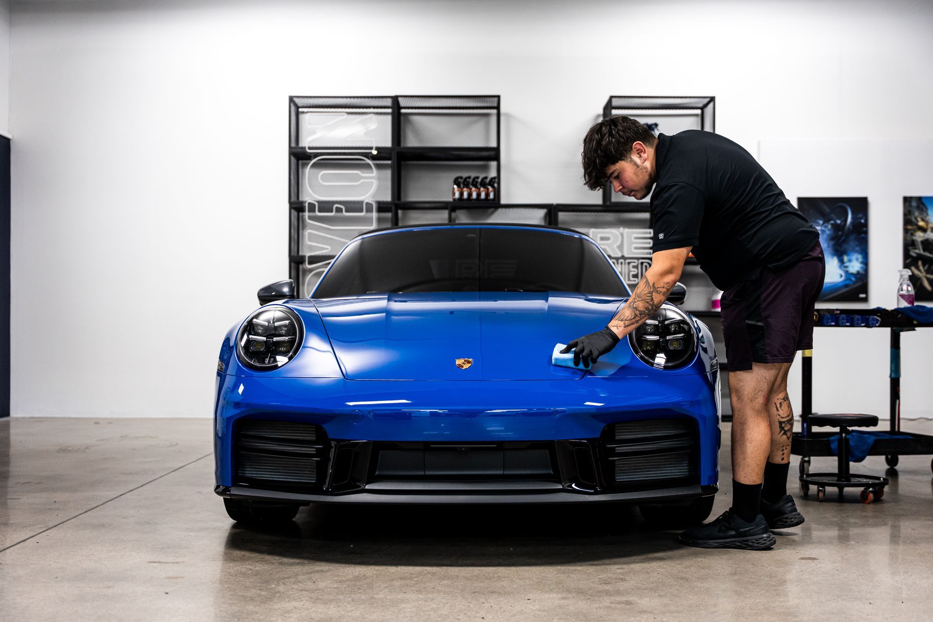Man detailing a blue Porsche in a well-lit shop, applying a coating to the front bumper.