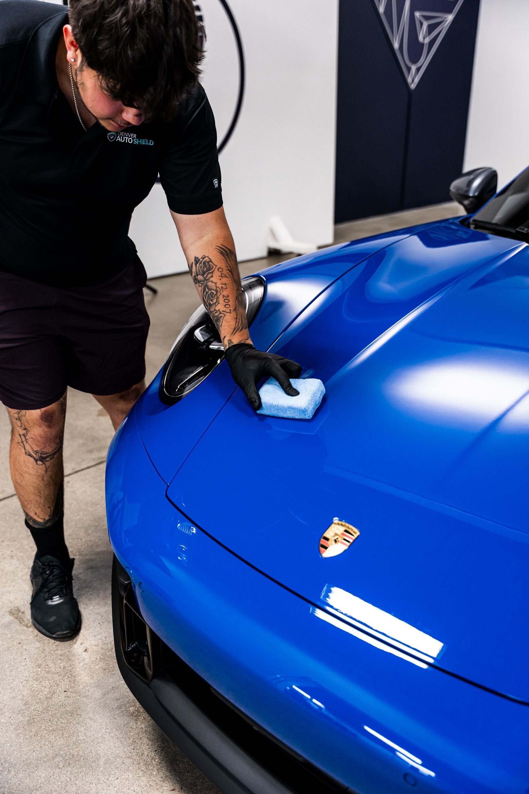 Man in black shirt and shorts detailing a bright blue Porsche, applying product with a sponge.