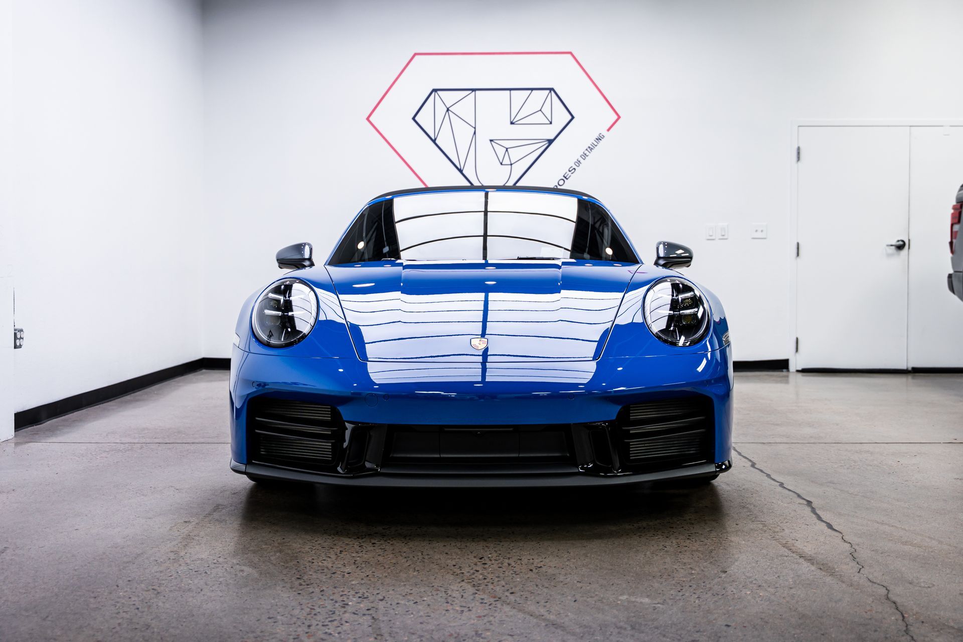 Blue Porsche 911 parked in a white garage; a geometric logo is on the wall.