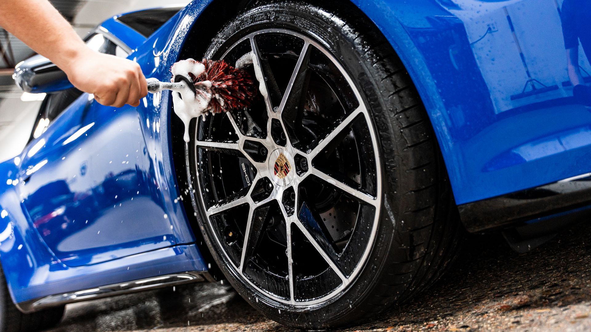 Person washing a blue sports car's wheel with a brush and soapy water.