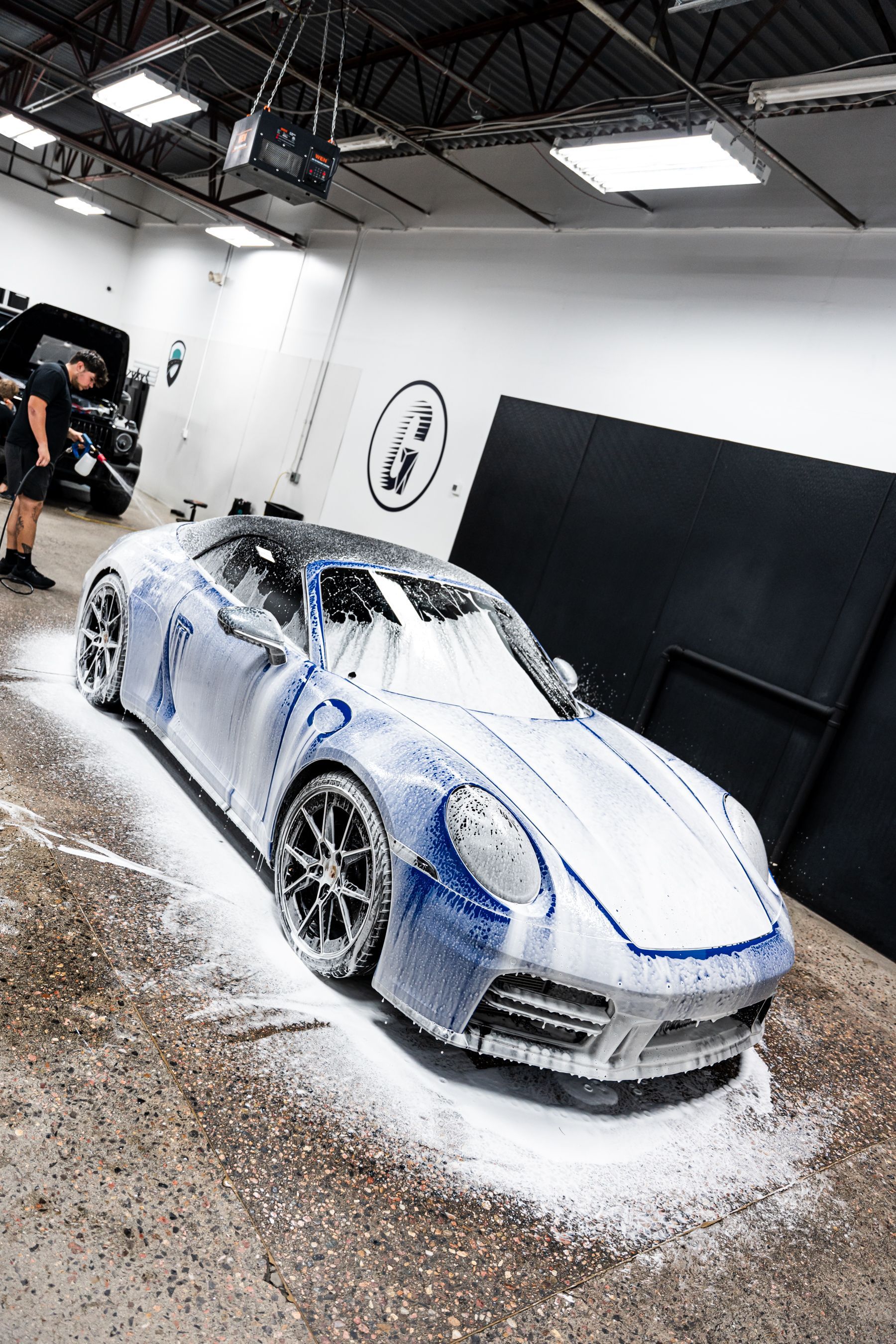 A blue sports car covered in white foam at a car wash facility. A person is nearby, holding a tool.