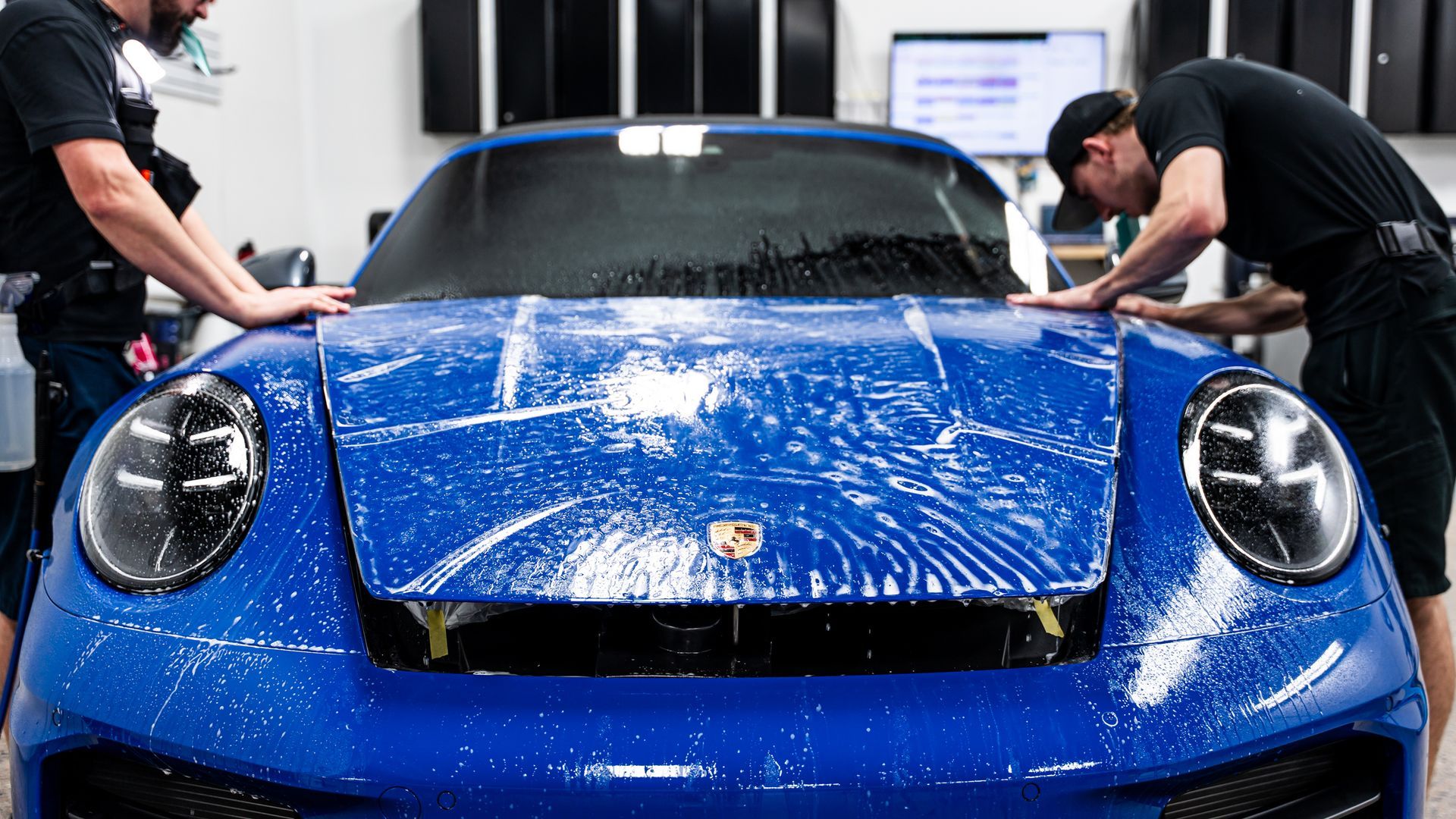 Two workers applying protective film to a blue Porsche in a shop.