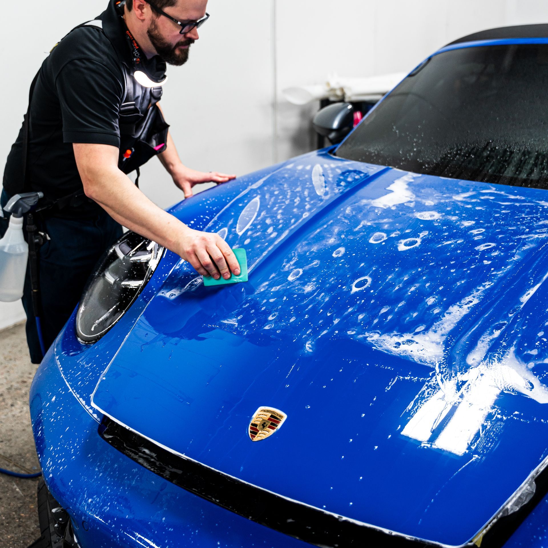 Man washing a bright blue Porsche car with a sponge, in a garage.