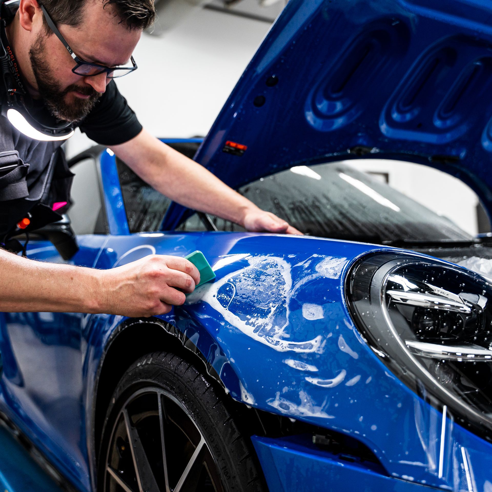 Man applies clear film to a blue car's fender, using a squeegee to remove bubbles.