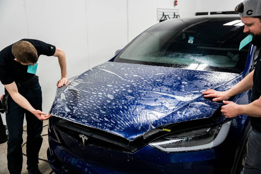 Two men are wrapping a blue tesla model x with a protective film.