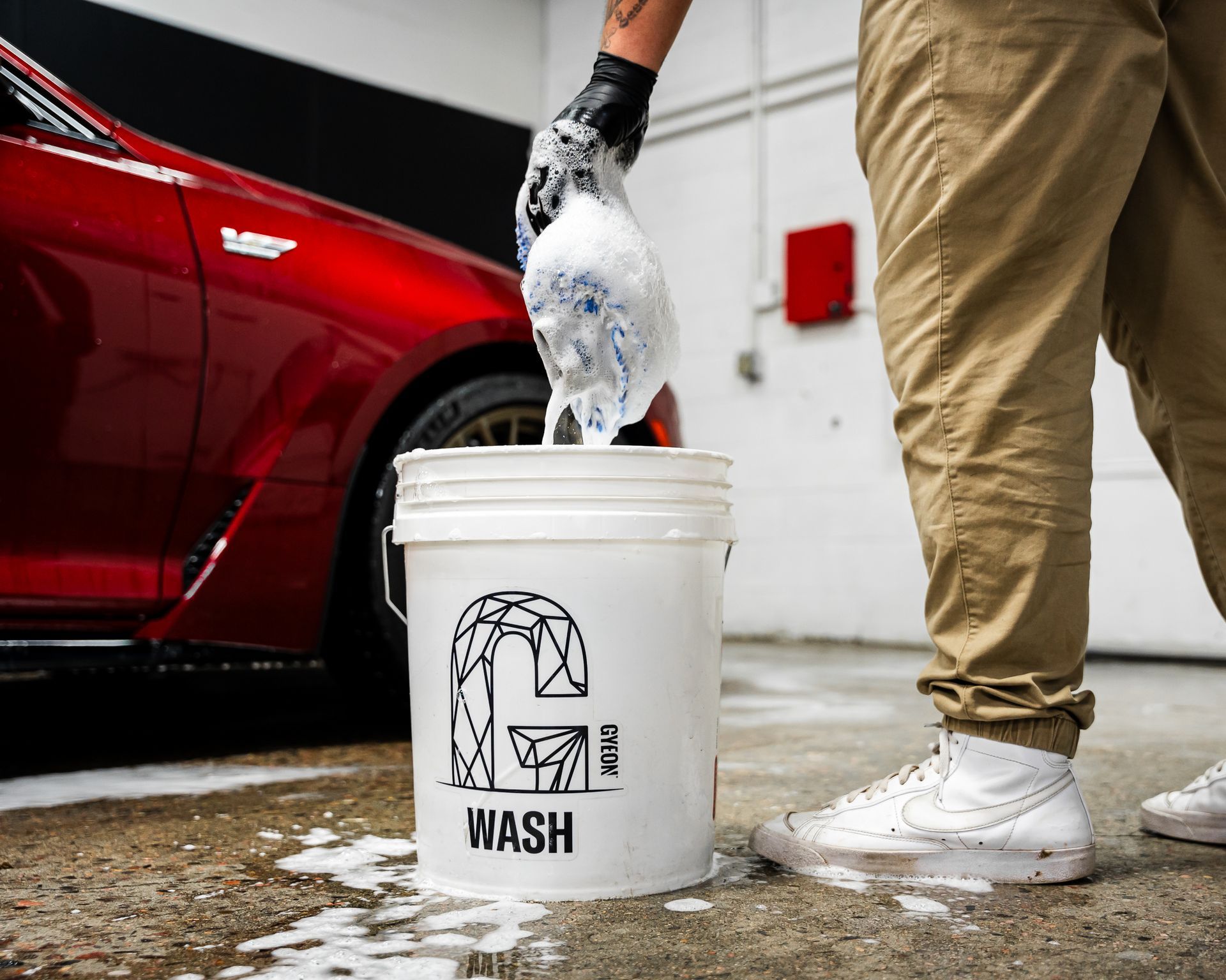 A person is pouring soap into a bucket in front of a red car.