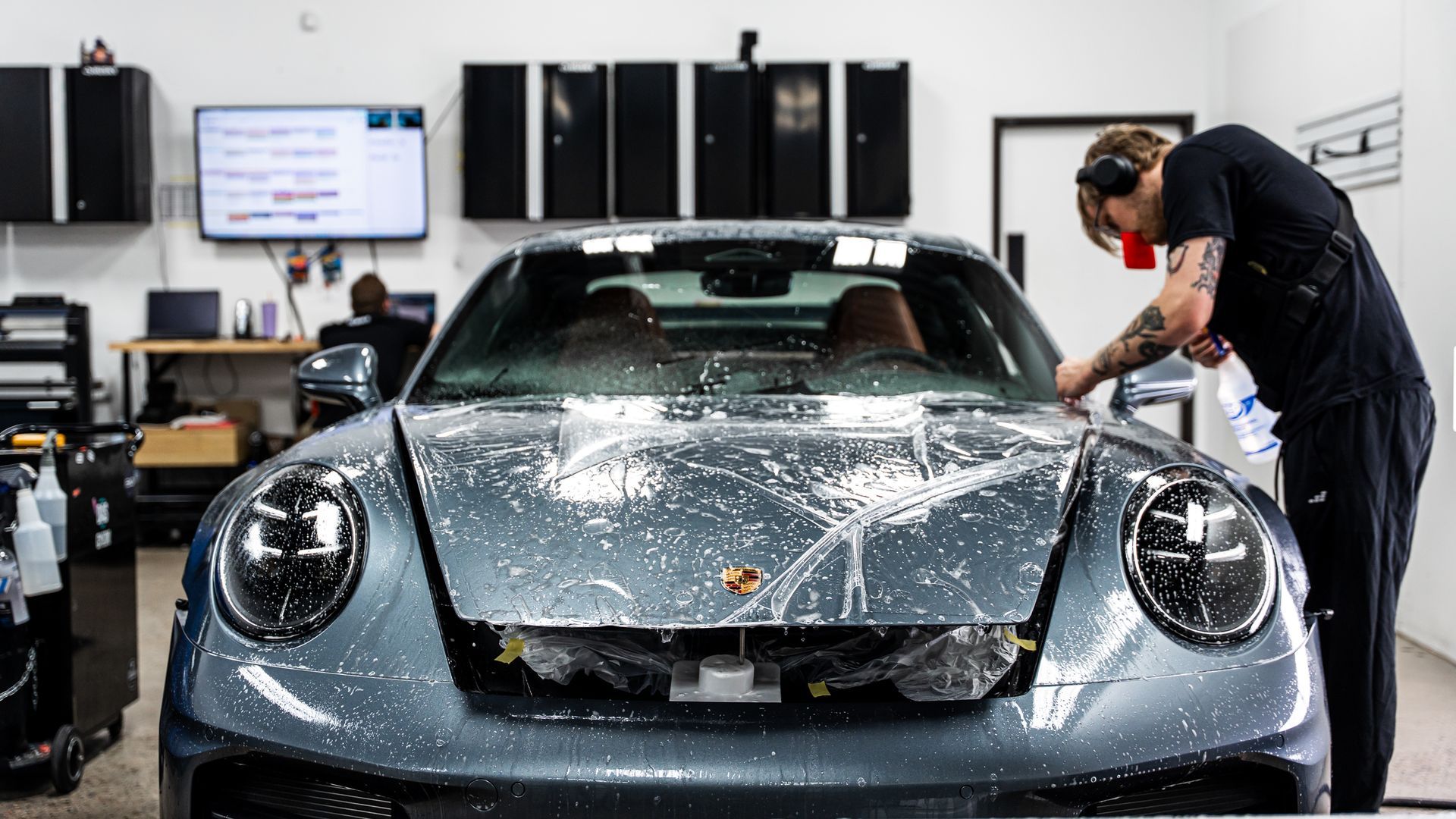 Person polishing a silver sports car in a garage, with equipment and monitors in the background