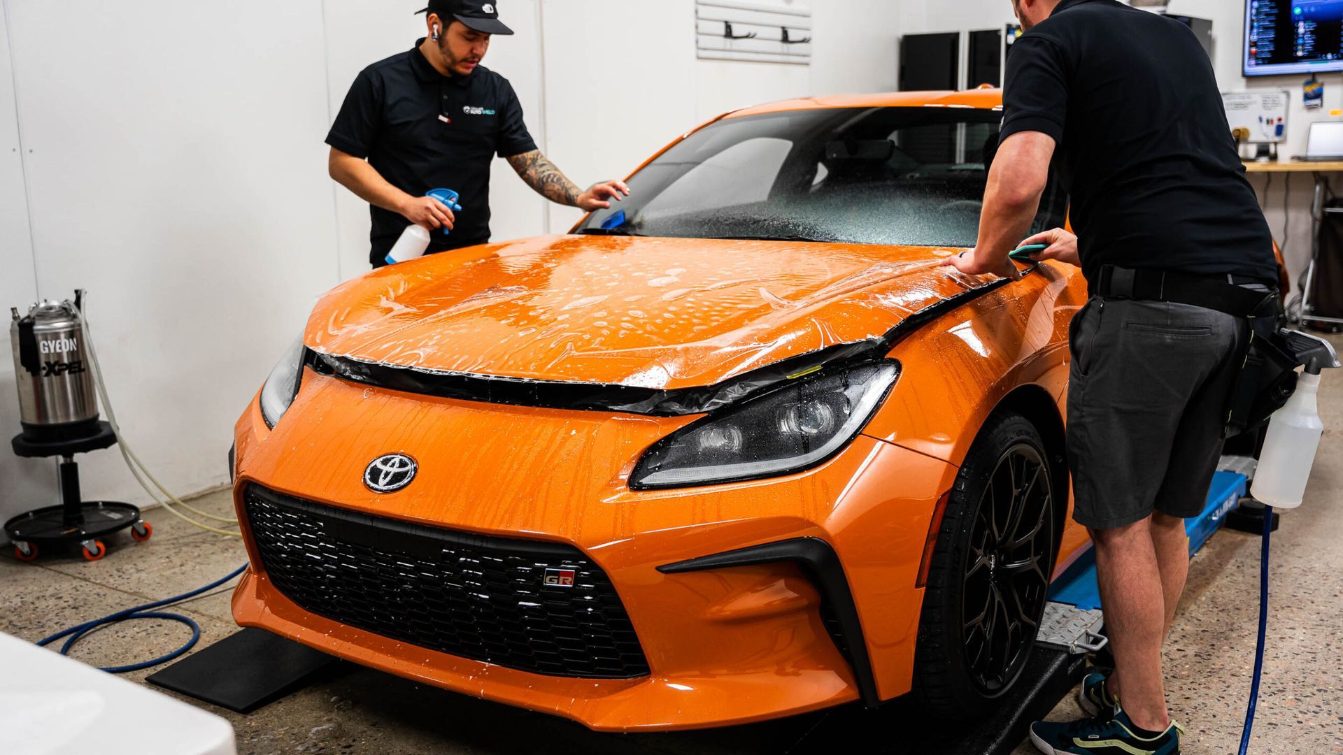 Two men are working on an orange car in a garage.