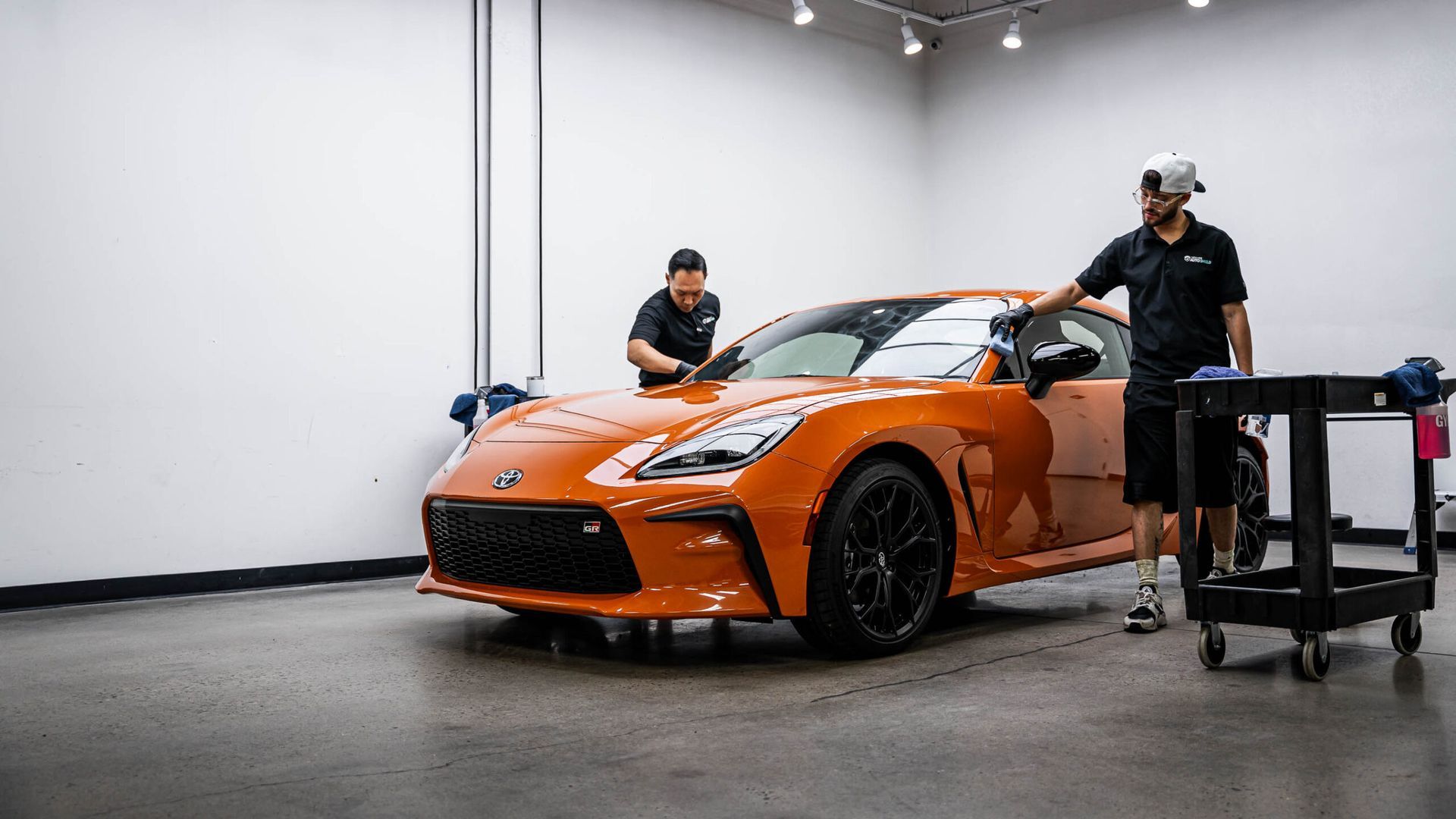 Two men are working on an orange sports car in a garage.