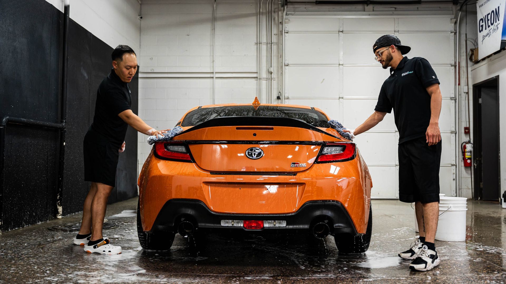 Two men are washing an orange car in a garage.