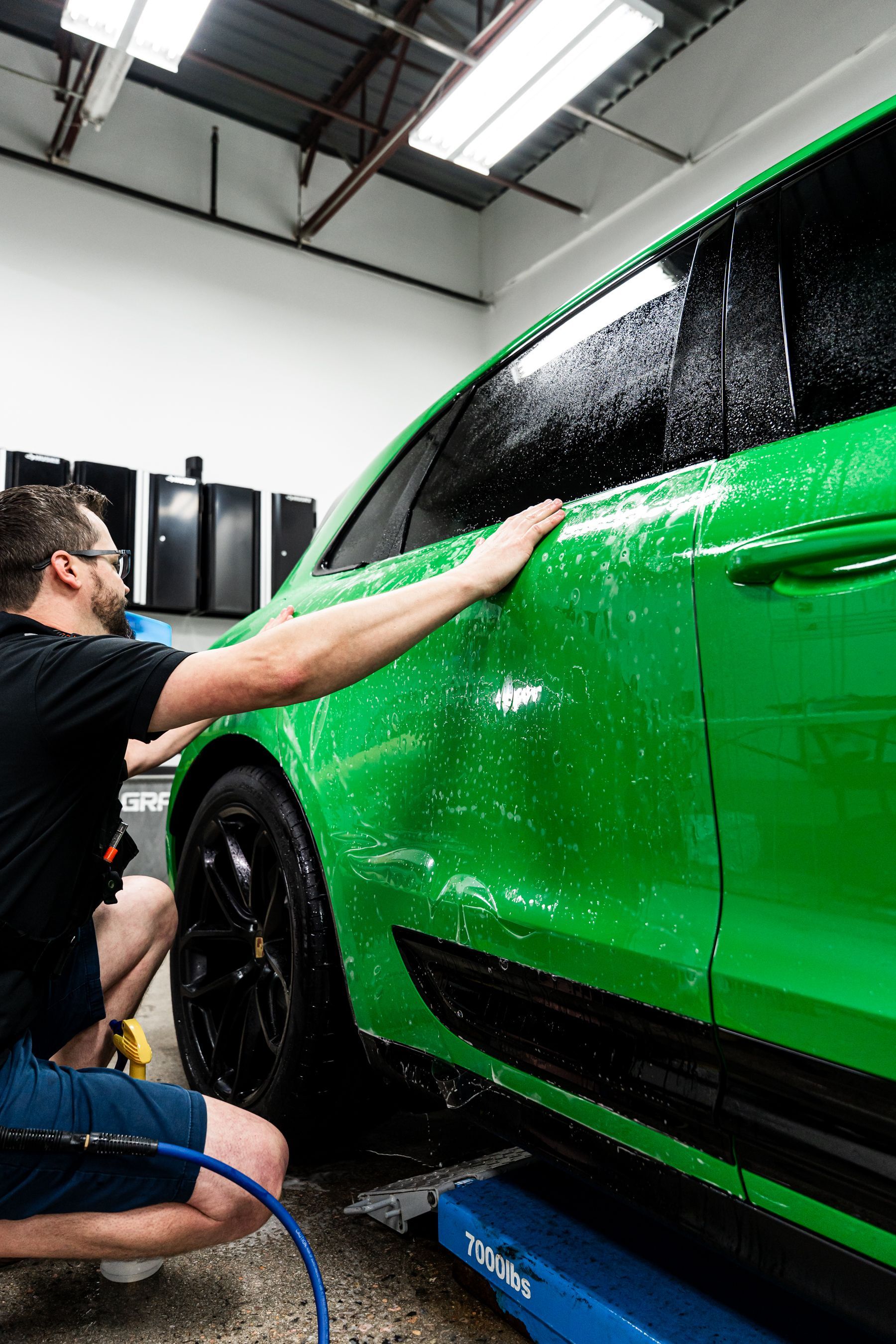 A person washing a bright green car, applying soap and water, inside a garage.