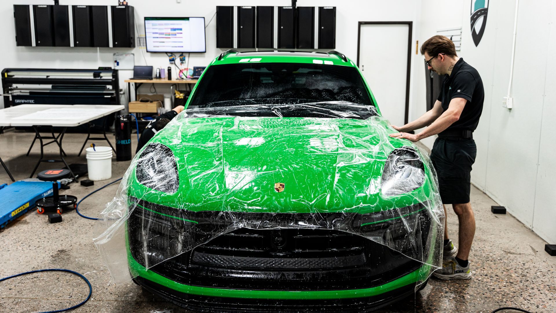 A person applying green protective film to the front of a green Porsche Macan in a shop.