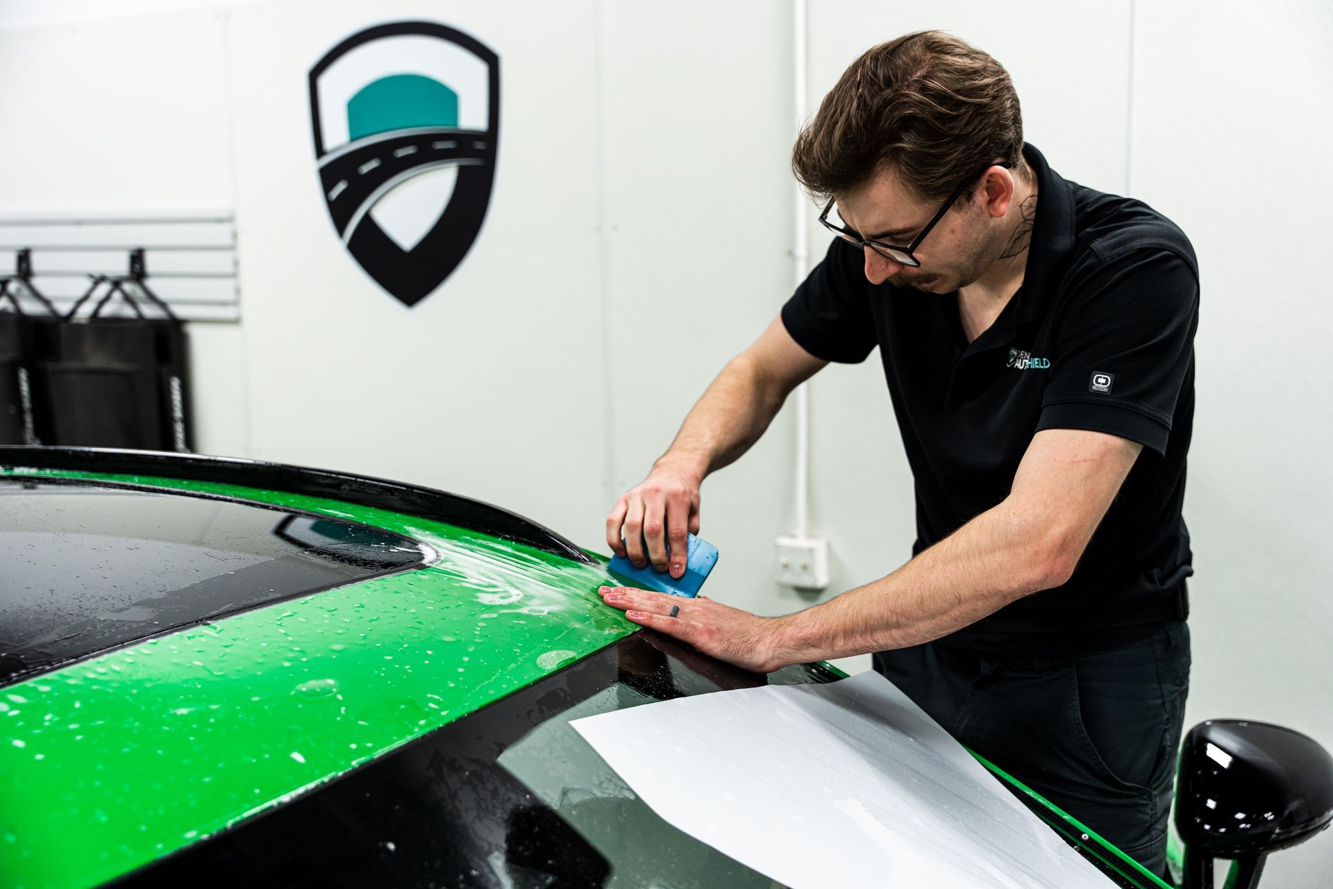 Man applying film to a green car roof, inside a shop. Logo on wall.