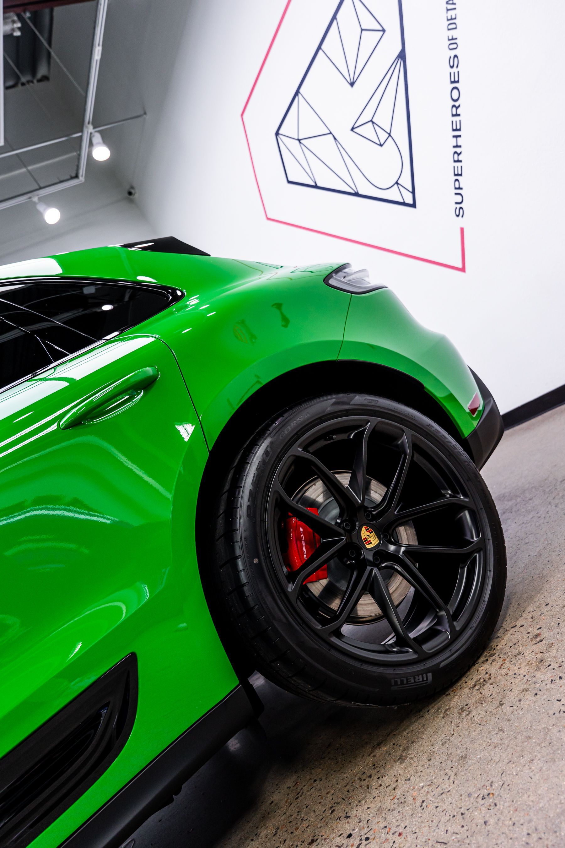 Green Porsche car with black wheels and red brake calipers parked indoors near white wall with geometric design.