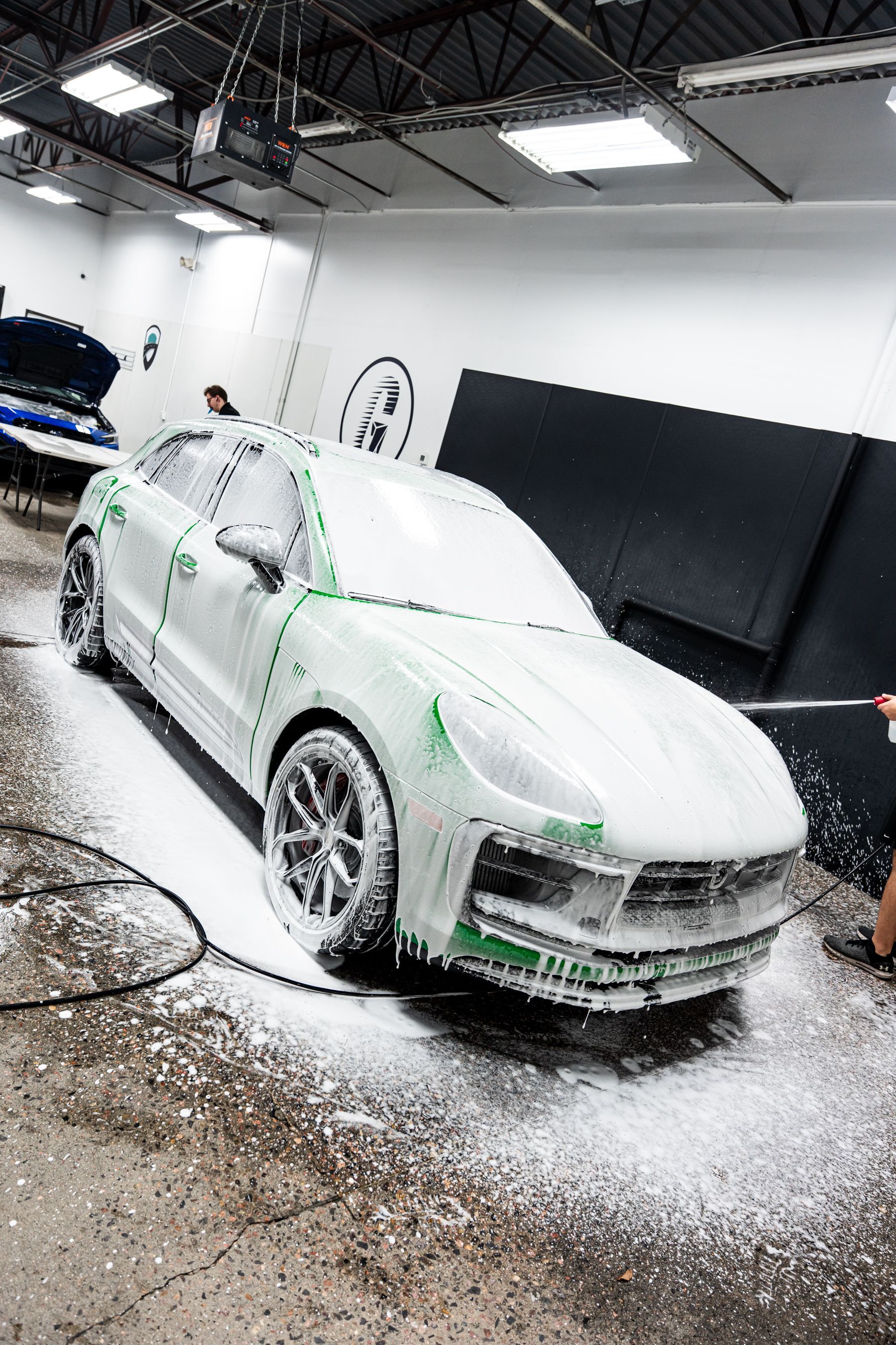 A white car covered in foam being washed indoors. A person is holding a hose.