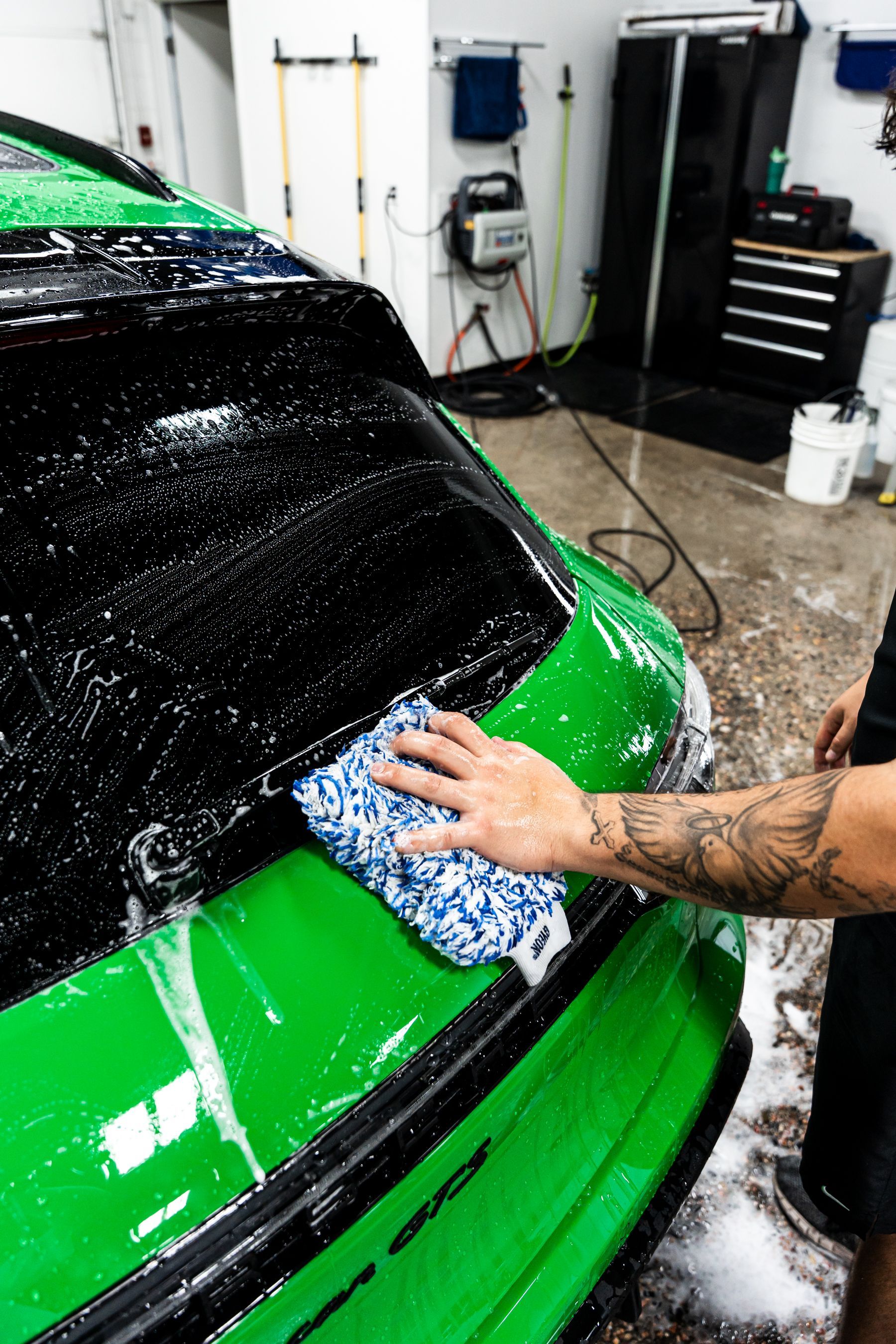 A person washing a bright green car with a soapy sponge in a garage.