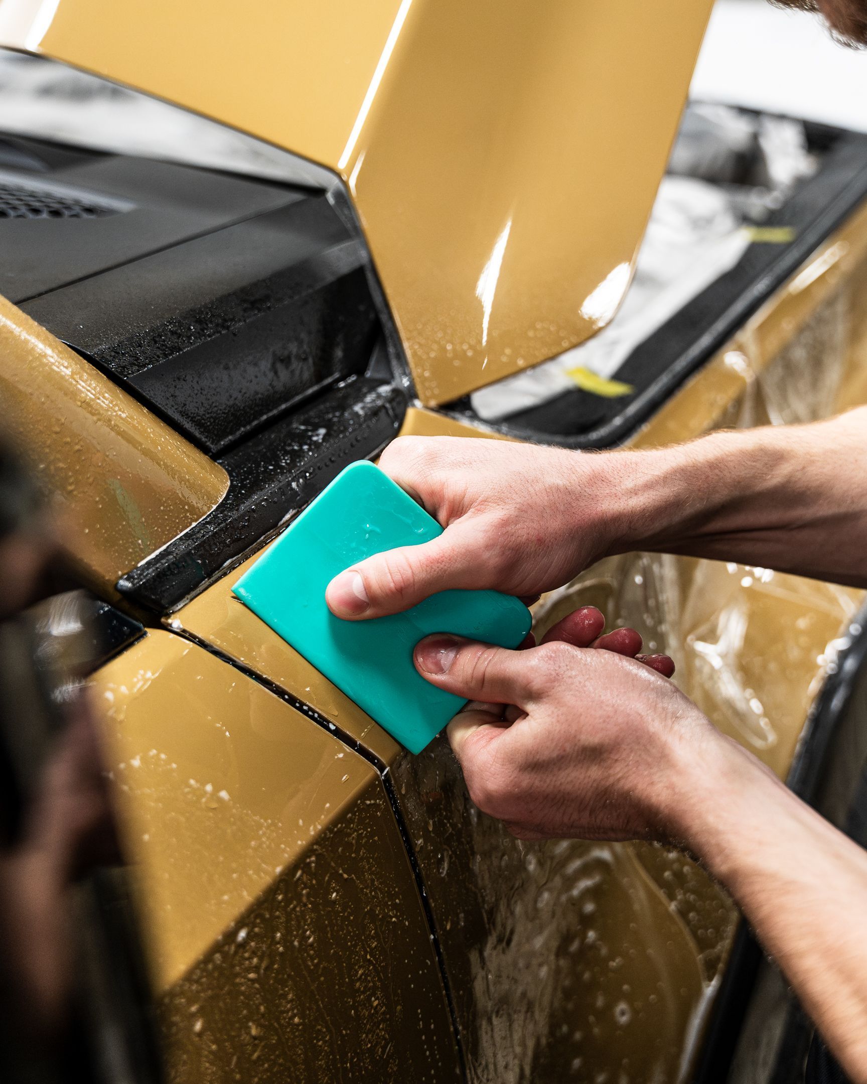 Hands using a teal squeegee to apply gold film to a car's body.