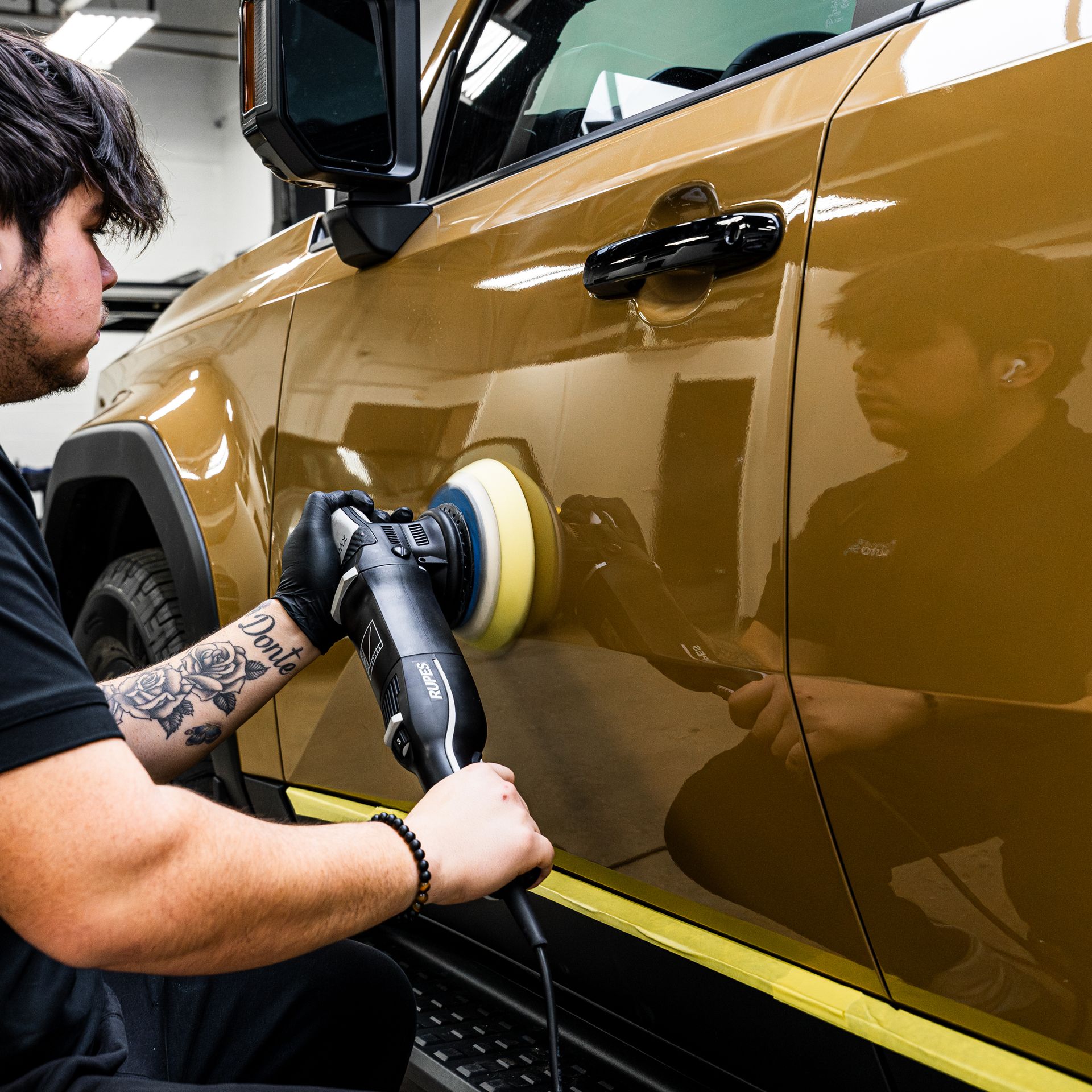 Man polishing a gold car door with an electric buffer in a garage.